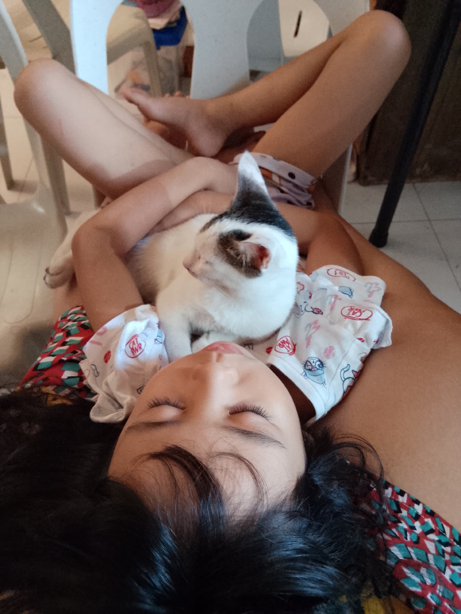The image captures a serene indoor scene featuring a young child, likely a girl, resting with a cat on her chest. The child lies on her back with closed eyes, suggesting she is asleep or deeply relaxed. She has long, dark hair spread around her head and wears a colorful, patterned top with green, red, and white geometric designs. A white piece of clothing, possibly a baby bib or a patterned cloth with cartoon animal motifs and text like "CAT PWR" (Cat Power) and "MEOW", covers her chest and abdomen, acting as a soft resting spot for the cat.
The cat is predominantly white with distinct black and brown markings on its head and back. It appears calm and is also resting, nestled against the child. The child's bare legs are visible in the upper background, bent at the knees, indicating a comfortable, casual posture. The setting appears to be a home in Davao City, Philippines, with hints of a tiled floor and furniture like white plastic chairs and a dark metal stand or chair in the background. The natural lighting suggests it is daytime. The overall impression is one of peaceful companionship and affection between the child and her feline friend.