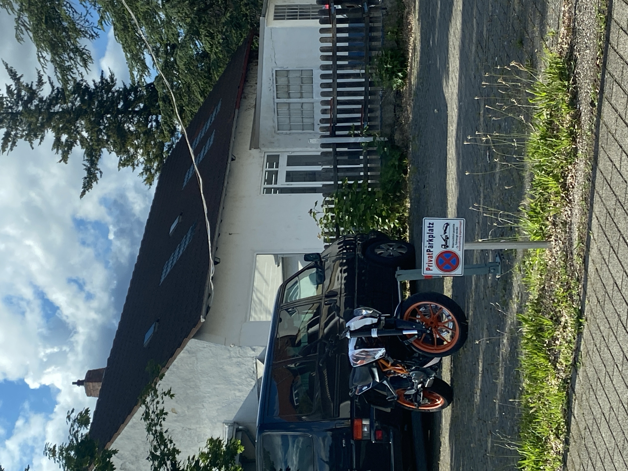 The image shows a black van parked on a private parking space (“Privatparkplatz”) in Landau in der Pfalz, Germany. A black and orange motorcycle is parked directly in front of the van. A white house with a dark roof and a small fence is in the background. There is vegetation to the right of the parking space, and trees and cloudy sky are visible in the background.