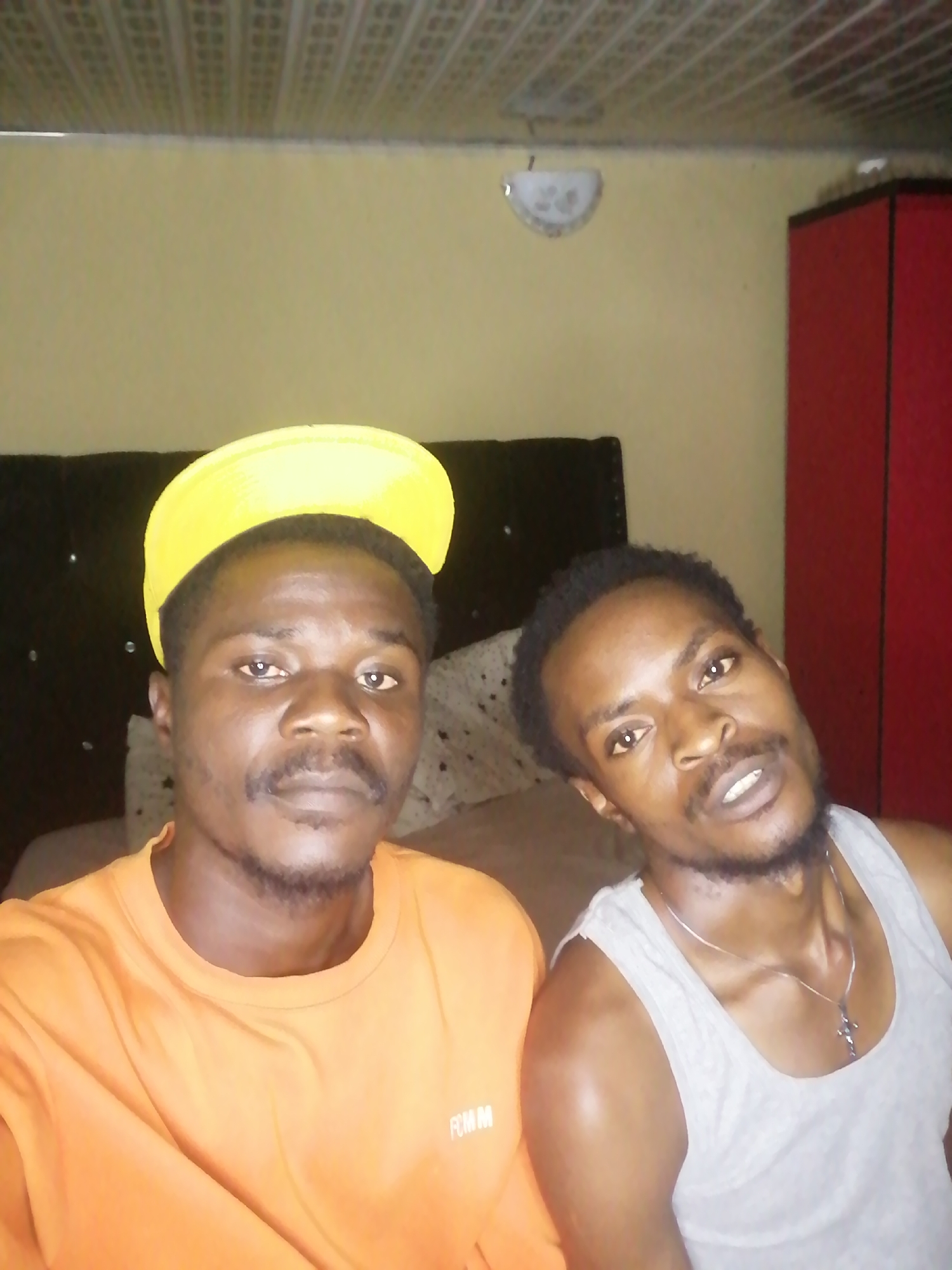 Two men are in an indoor setting. The man on the left is wearing an orange shirt and a yellow cap. The man on the right is wearing a gray tank top. They are both looking at the camera. Behind them is a black headboard, a light yellow wall, a ceiling with square panels, and a red cabinet. The location is Unknown, Unknown.