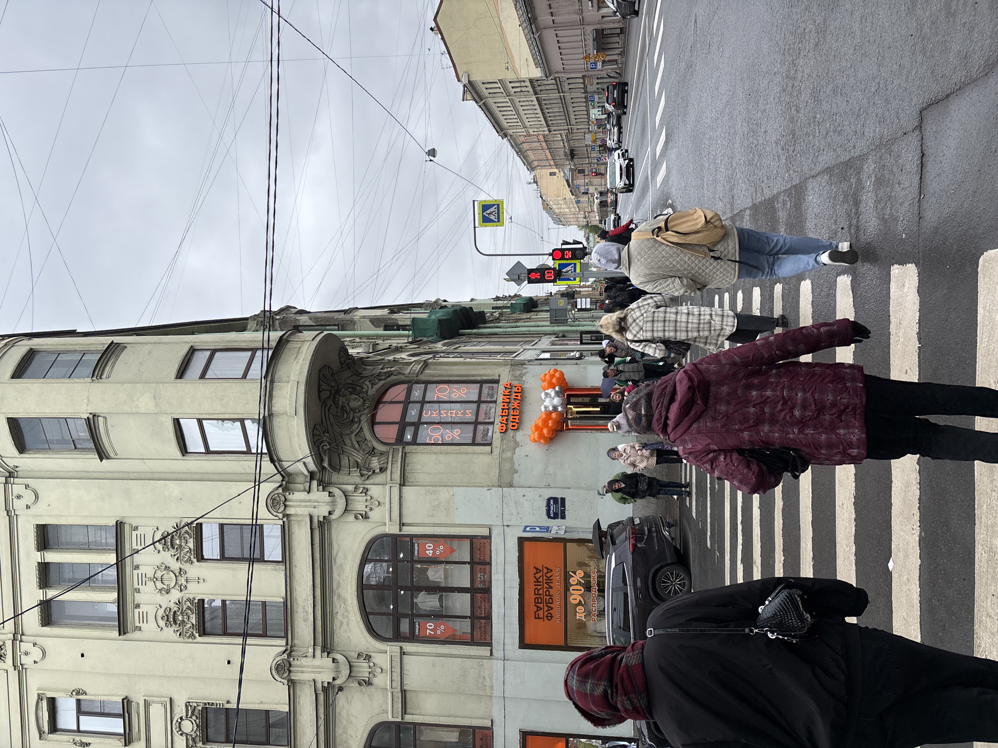 The image shows a street scene in Saint Petersburg, Russia. Pedestrians are crossing a zebra-striped crosswalk. A building with ornate architectural details and windows displays advertisements and discounts, with balloons marking the entrance to a store called "Fabrika Odezhdy" (Clothing Factory) and "Fabrika Furnitury" (Furniture Factory). Several people are seen on the sidewalk and street, with cars parked and passing by. A traffic light in the distance shows a red light. Overhead wires crisscross the sky. The sky is overcast. The building’s sign has text in Cyrillic script.
