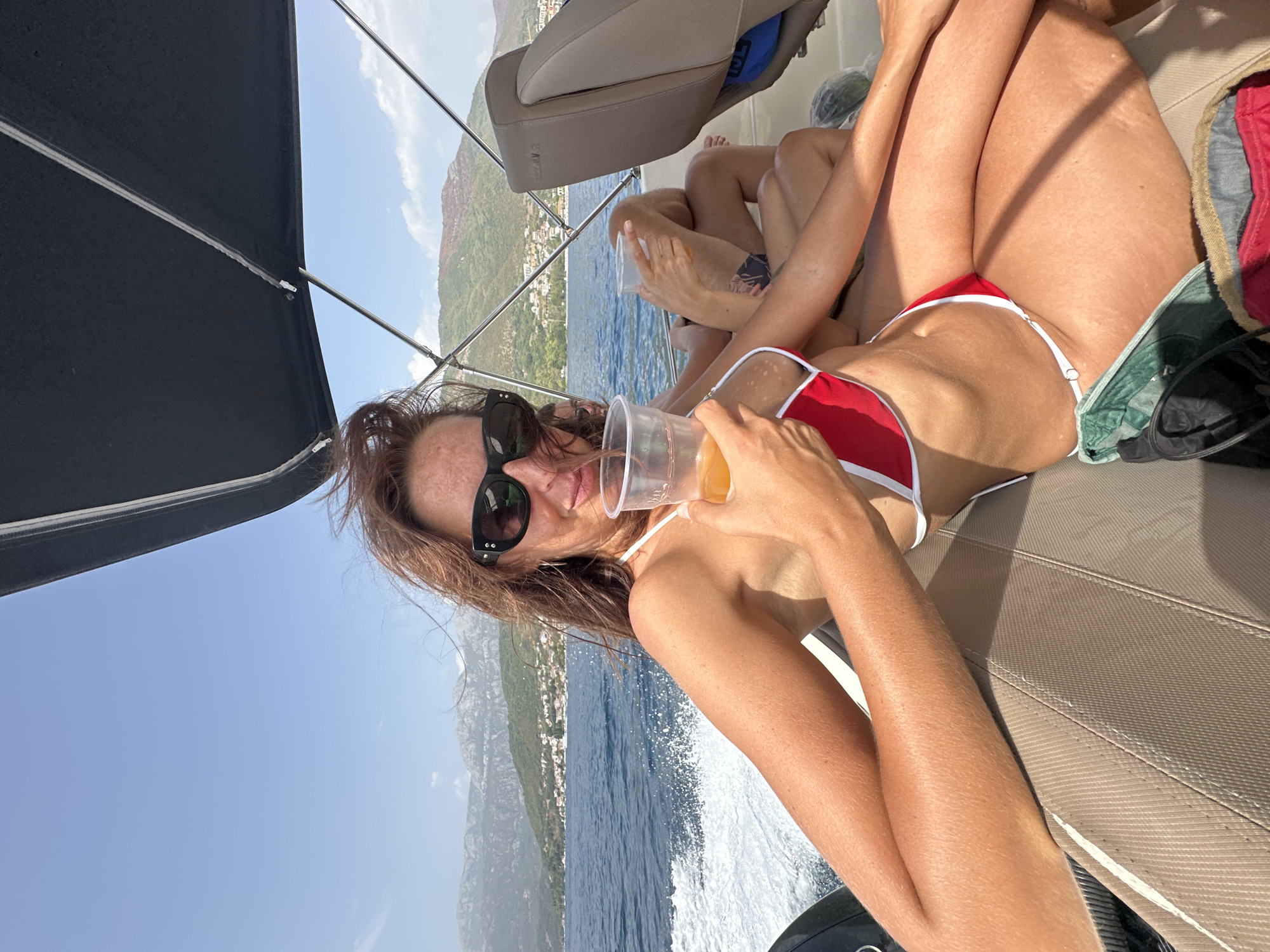 A woman with auburn hair, wearing black sunglasses and a red and white bikini, sits on a boat in the waters off Kumbor, Montenegro. She holds a plastic cup of orange liquid in her right hand. Her left arm rests on her lap. Behind her, the water is choppy and churns with white foam. Mountains covered in green vegetation and dotted with buildings rise in the background, under a clear blue sky. Another person is partially visible sitting behind her, holding a similar drink. The boat’s interior, with upholstered seating, is visible.