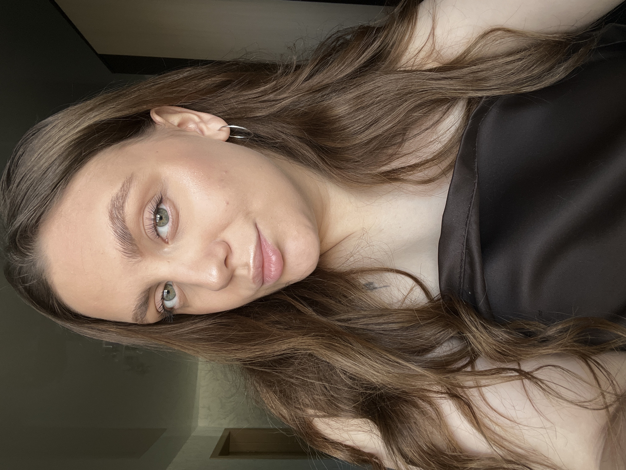 A young woman with fair skin, long brown hair, and green eyes is posing for a selfie. She is wearing a dark, satin-like top and a silver hoop earring in her left ear. The background appears to be an interior space with a wall and a glimpse of a door. There's a small tattoo visible near her collarbone. The image does not definitively place the location in Chaikovsky, Russia.