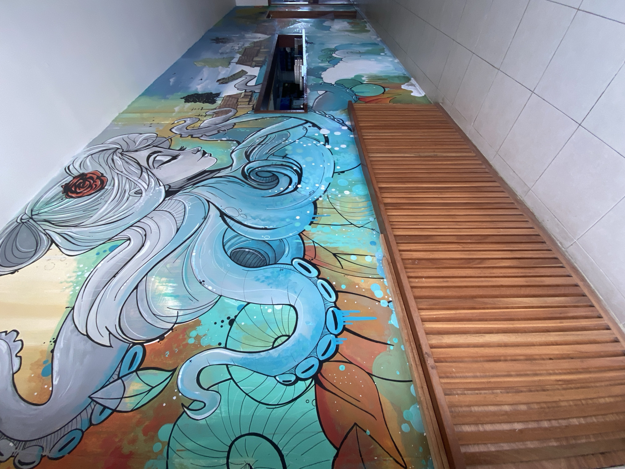 The image shows a hallway in Baan Chaloklum, Thailand. The left wall is decorated with a vibrant mural depicting a stylized woman with an octopus. The mural includes various colors and patterns, with a blue octopus dominating the scene. Above the mural, near the top of the wall, is a rectangular mirror or window. To the right of the mural is a set of horizontal wooden slats, possibly part of a folding screen or door. The right wall is covered with white square tiles. The perspective suggests the photo was taken looking upwards along the hallway.