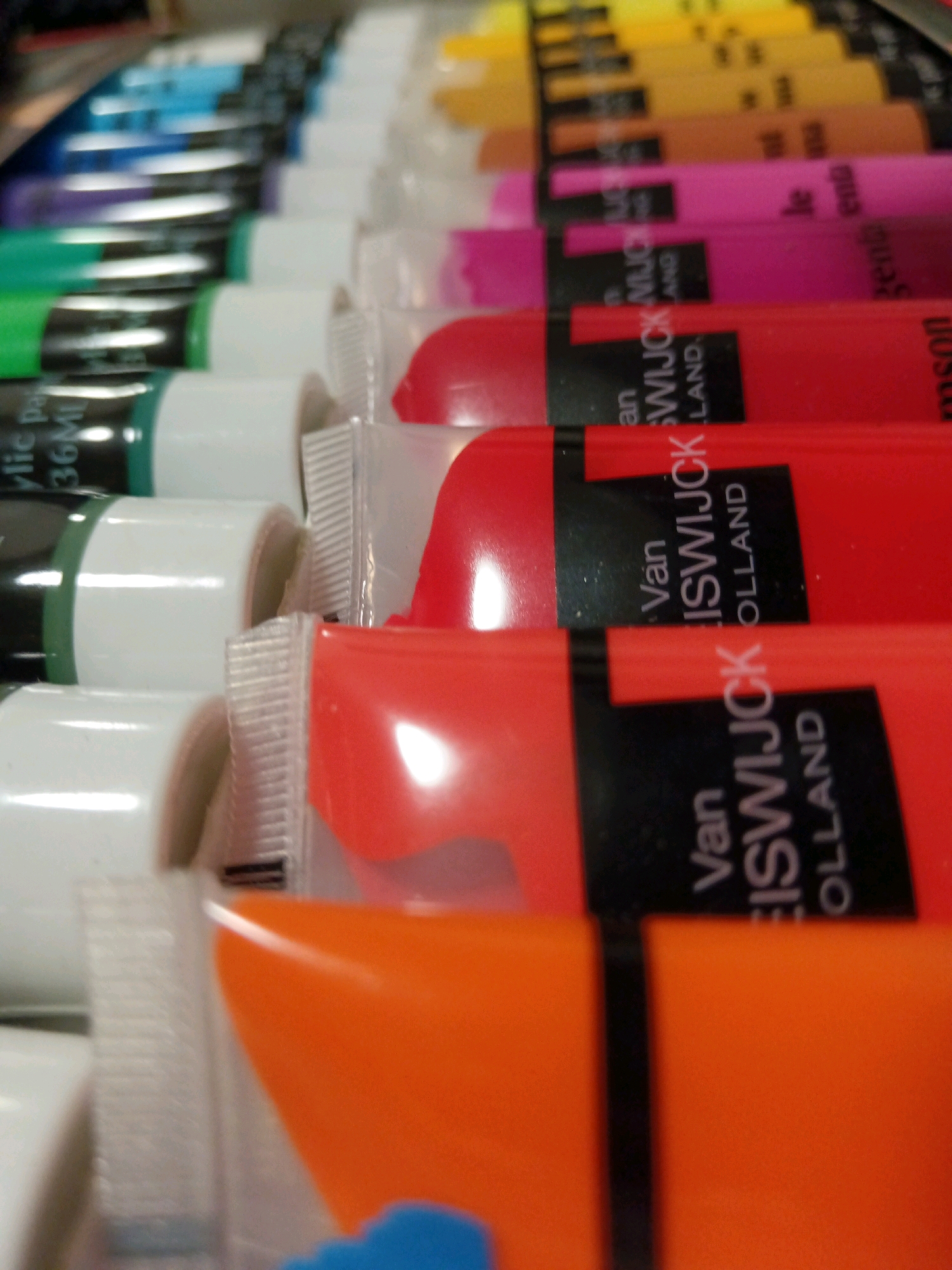The image displays a close-up, angled view of several tubes of paint arranged in rows. The tubes are varied in color, including green, red, pink, and yellow. Some tubes have black labels with white text, indicating brands or contents. The lighting is bright, creating reflections on the plastic surfaces of the tubes. The background is blurred and indistinct, suggesting a shallow depth of field. There are no people, animals, or identifiable landmarks visible in the image. The scene appears to be an indoor setting, possibly a retail display or a personal art supply collection. No specific location in Kempten (Allgäu), Germany, can be determined from the visual information provided.