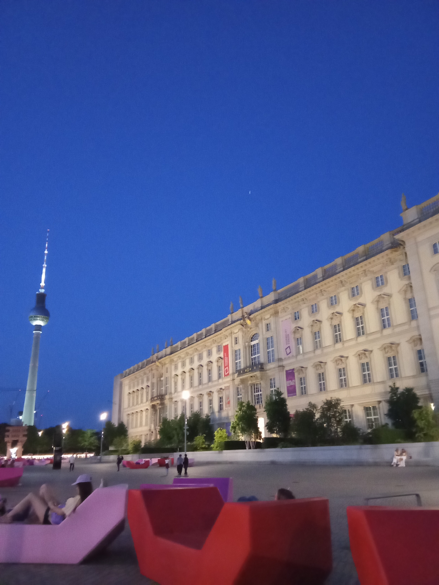 The image shows an outdoor scene in Berlin, Germany, at dusk or night. In the background, the Fernsehturm Berlin television tower is visible on the left, and a large, ornate, light-colored building is on the right, likely the Berlin City Palace. The sky is a deep blue.
In the foreground, there are several brightly colored, modern-looking lounge chairs in pink and red hues. People are seen relaxing in some of these chairs. In the middle ground, figures of people are walking across a paved area. Some trees are present between the building and the paved area. Street lights are also visible.