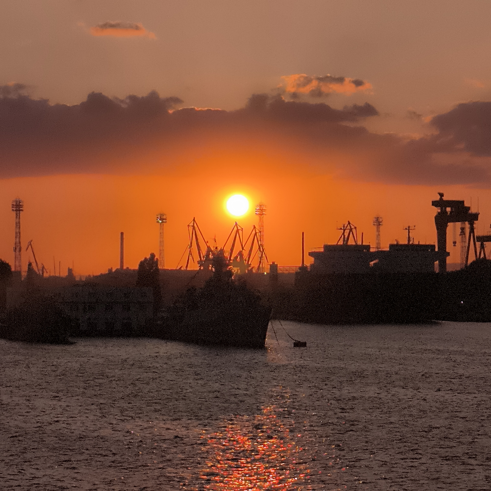 The image depicts a sunset over a port area in Varna, Bulgaria. The sky is filled with warm hues of orange and red, with scattered clouds. The sun, a bright, luminous orb, is positioned slightly to the left of the center of the frame. Silhouetted against the vibrant sky are several large cranes and industrial structures, characteristic of a harbor. Two large ships are visible, one in the foreground and another further back, their dark forms contrasting with the illuminated sky. The water in the foreground reflects the fiery colors of the sunset, creating a shimmering, almost molten effect. The overall scene is one of industrial activity set against a dramatic natural backdrop.