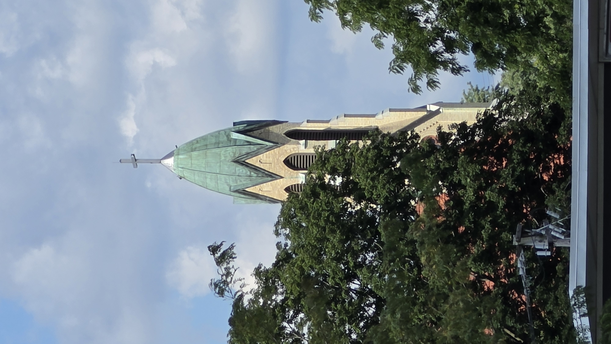 The image shows the upper portion of a church steeple in McHenry, United States of America. The steeple is constructed of light-colored brick and features a green, conical roof topped with a cross. The steeple is partially obscured by lush green trees. The sky is partly cloudy with scattered white clouds against a blue backdrop. Below the steeple, glimpses of red roofing tiles can be seen through the foliage, along with parts of a telephone pole with multiple insulators and wires. The lower edge of the image shows a portion of a white overhang or structure.