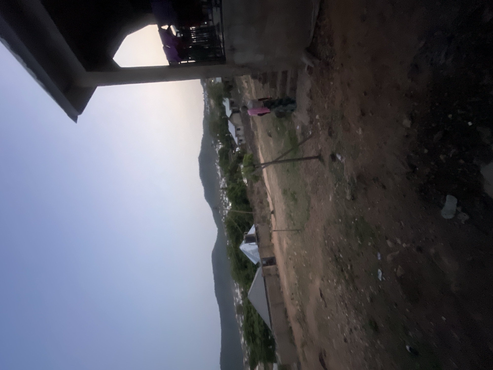 The image depicts a view from an elevated position in Lokoja, Nigeria. A building structure is visible in the foreground with clothes hanging on a balcony. Beyond the building, a landscape unfolds with scattered houses, vegetation, and a mountain range in the distance. The sky is transitioning in color, indicating either sunrise or sunset. A person stands in the middle ground, close to a clothesline. The ground is primarily dirt and sparse vegetation.