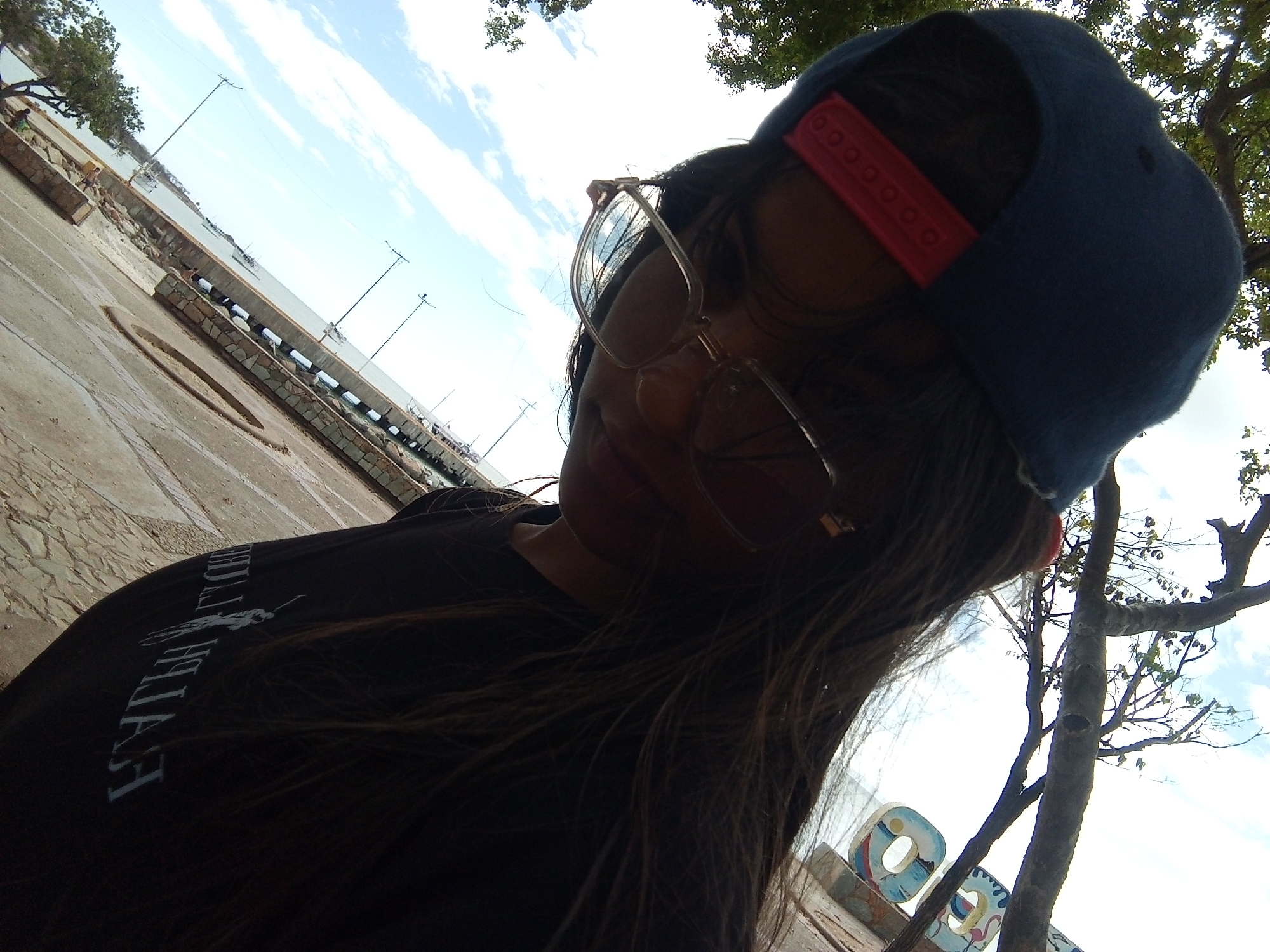 The photograph shows a woman with long brown hair wearing a blue baseball cap with a red adjustable strap and glasses. She is wearing a dark-colored t-shirt with white lettering. The photograph is taken from a low angle, looking up at her. In the background, there is a view of what appears to be the coastline of Juan Griego, Venezuela, visible with water, a pier-like structure, and street lights. Part of a painted sign can also be seen in the lower right corner. A tree with sparse leaves is visible in the upper right corner of the frame. The surface in the foreground has cracks and variations in texture suggesting it might be concrete or pavement. The sky is light blue with some clouds.