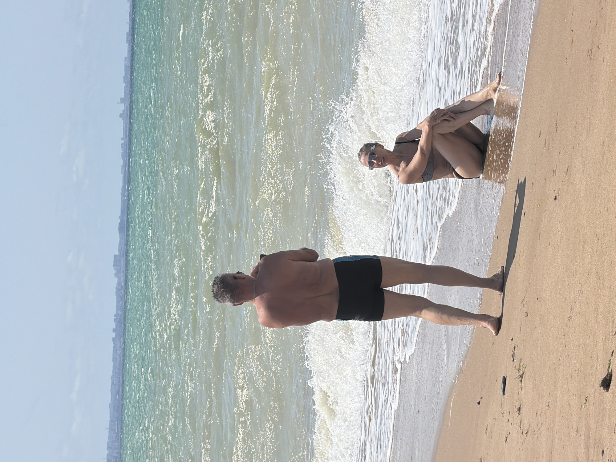The image shows a man and a woman on a sandy beach with the sea in the background. The man, seen from the back, is wearing black swim trunks and holding a phone, possibly taking a photo. He has short, graying hair. The woman is seated in the shallow water near the shore, facing left, wearing a dark bikini and sunglasses. She is resting her arms on her knees. The sea is a vibrant turquoise with white-capped waves. In the far distance, a city skyline with buildings is visible across the water. The sky is bright and clear. The location is identified as the Качинский округ, Ukraine.