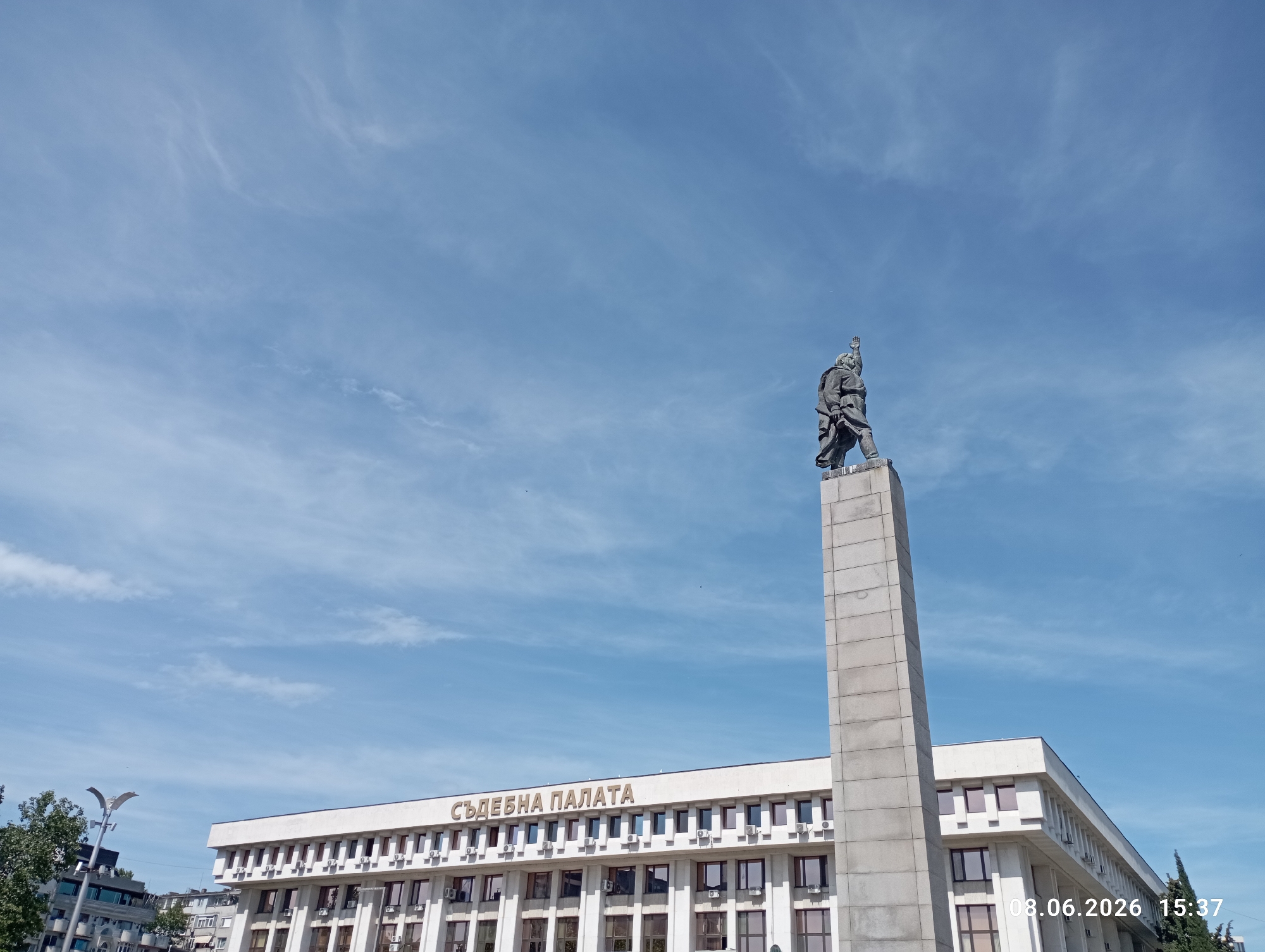 The image shows a wide-angle, low-angle shot of the Palace of Justice (Sŭdebnata Palata) in Burgas, Bulgaria, under a bright blue sky with scattered clouds. A large concrete obelisk with a statue of a man in uniform stands to the right of the frame, reaching towards the sky. The statue is depicted with his right arm raised. The Palace of Justice building is a long, white, modern structure with multiple windows and air conditioning units visible. The words "СЪДЕБНА ПАЛАТА" (Palace of Justice) are written in gold lettering above the main entrance. The timestamp "08.06.2026 15:37" is visible in the bottom right corner of the image. The foreground shows the tops of trees and some streetlights. The overall scene is an outdoor urban environment on a clear day.