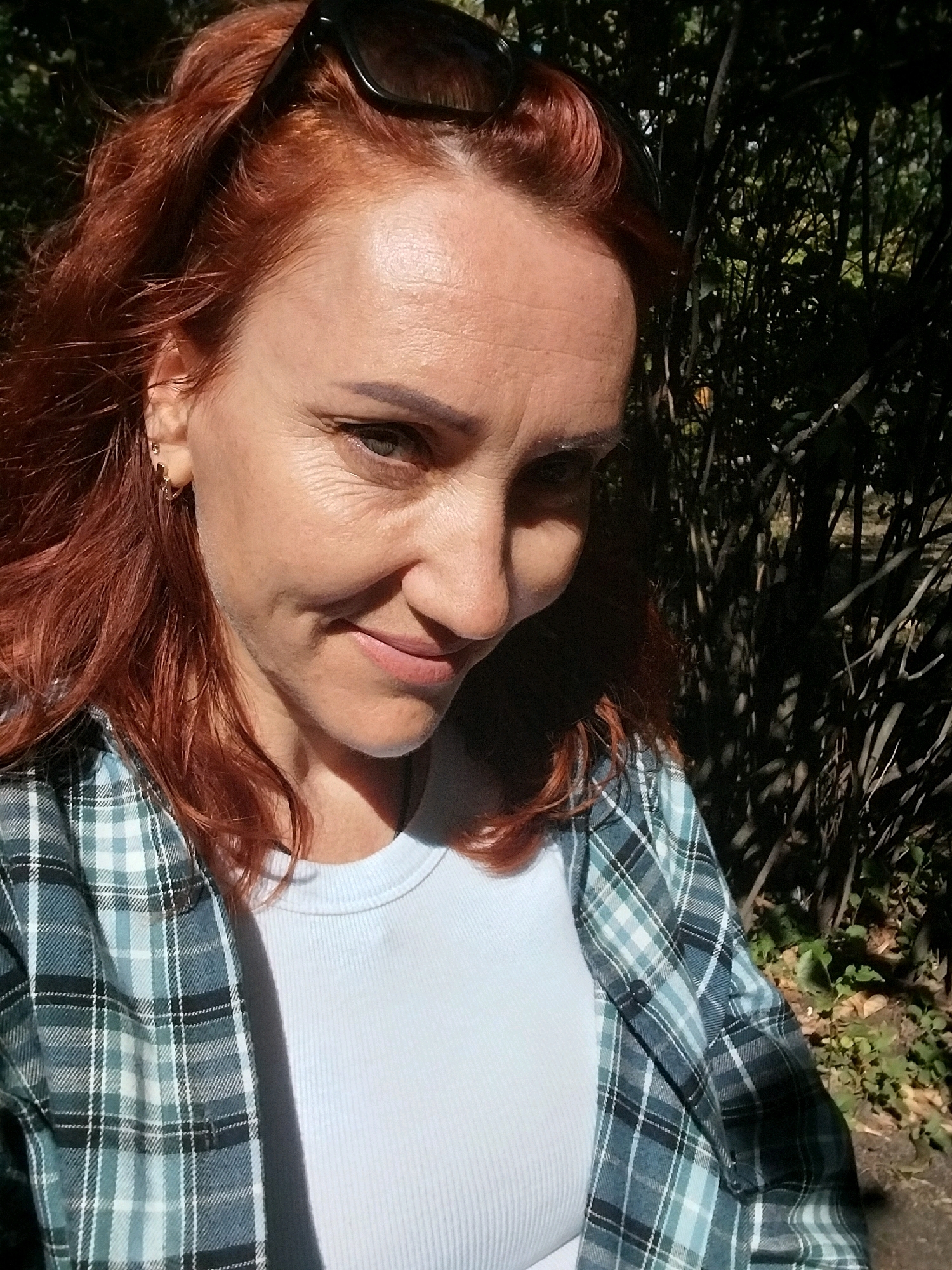 A medium shot shows a Caucasian woman with reddish-brown hair and fair skin, taken outdoors. She is wearing a light blue ribbed t-shirt and an open plaid shirt with blue, green, and white checkered patterns. She has on sunglasses perched on her head. The background consists of dense foliage and trees, suggesting a park or natural area. The lighting indicates it is daytime, with some shadows cast on the subject. There is no visible indication of the city of Dnipro, Ukraine in the image.