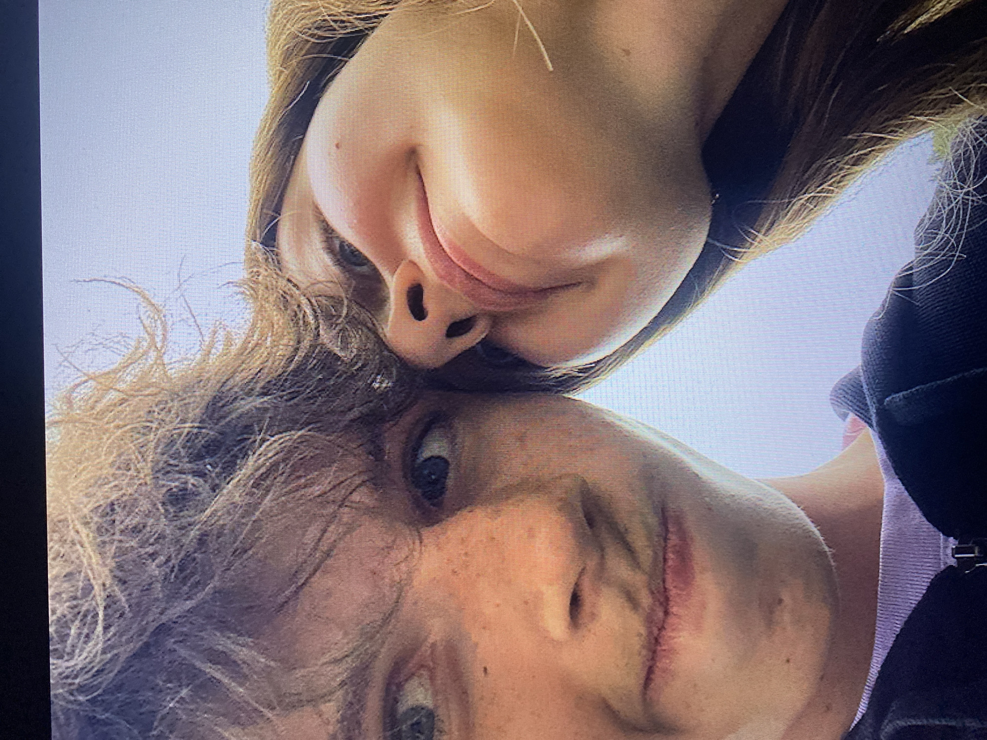 The image is a close-up, low-angle selfie featuring two individuals. On the left, a young person with fair skin, freckles, and messy brown hair is looking directly at the camera with a slightly amused expression, lips slightly parted. Their eyes are open wide. On the right, another person's face is partially visible, tilted upwards and away from the camera, with their eyes closed and a subtle smile. Their hair is long and brown, falling across their cheek. Both individuals appear to be outdoors, with a bright, overcast sky visible in the background. The foreground shows the upper chest area of the person on the left wearing a light purple t-shirt and a dark zipper jacket. The image quality is grainy, suggesting it was taken with a phone camera or a similar device. There are no identifiable landmarks that would allow for specific location identification in Poznan, Poland.