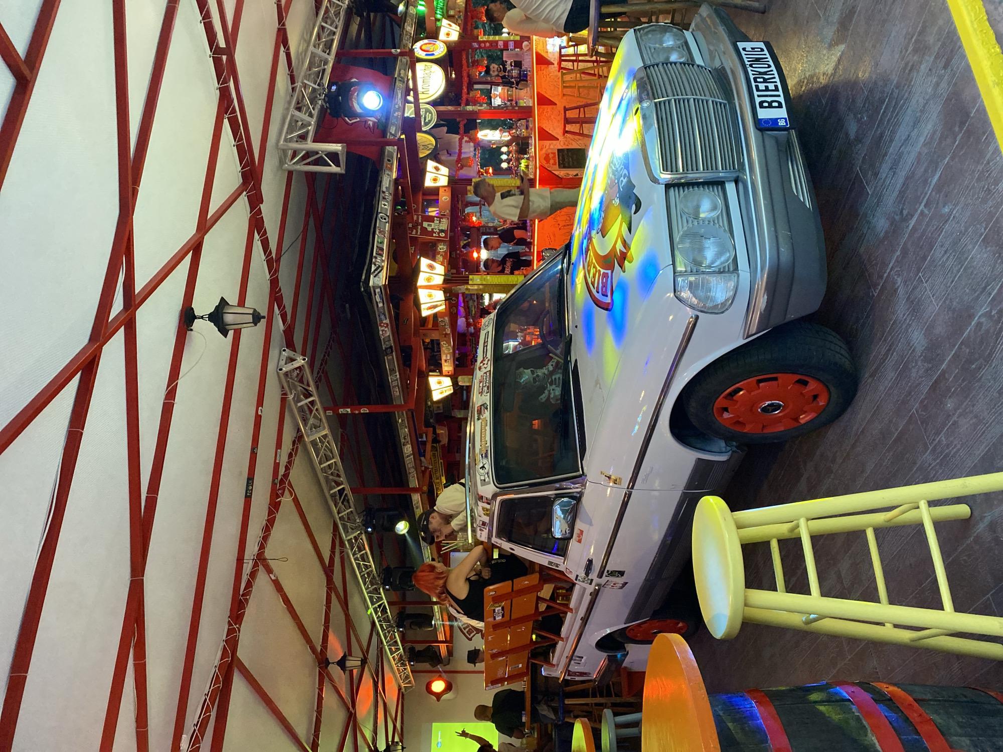 The image depicts an indoor entertainment venue, identified as a bar or club, located in Varna, Bulgaria. The ceiling consists of a white, semi-translucent material supported by a red metal beam framework, from which traditional black lanterns and modern stage lighting trusses with blue spotlights are suspended.
Central to the scene is a white Mercedes-Benz W123, notably modified into a pickup truck. It features vibrant red hubcaps and a colorful graphic design, predominantly blue and yellow, on its hood. The front license plate displays "BG BIERKONIG." The truck bed is fitted with two rows of wooden bench seating where a woman with red hair, wearing a black top, is engaged in conversation with a man in a light-colored shirt and a cap. A skull prop occupies the driver's seat.
In the background, a brightly lit bar counter spans the width of the venue, illuminated by a prominent orange-red glow. Several beer taps, branded beer signs (e.g., Krombacher), and merchandise are visible behind the counter. Multiple individuals, including bar staff, are present behind and in front of the bar. One man in a light shirt is seen looking at a mobile phone. Another man in the left background raises his arm.
The flooring is composed of dark wooden planks. Various furniture elements include dark wooden barrel-shaped high tables with bright orange circular tops, and tall, light yellow wooden stools. The overall environment suggests a lively, social atmosphere with a mix of ambient and direct lighting creating a dynamic visual experience.