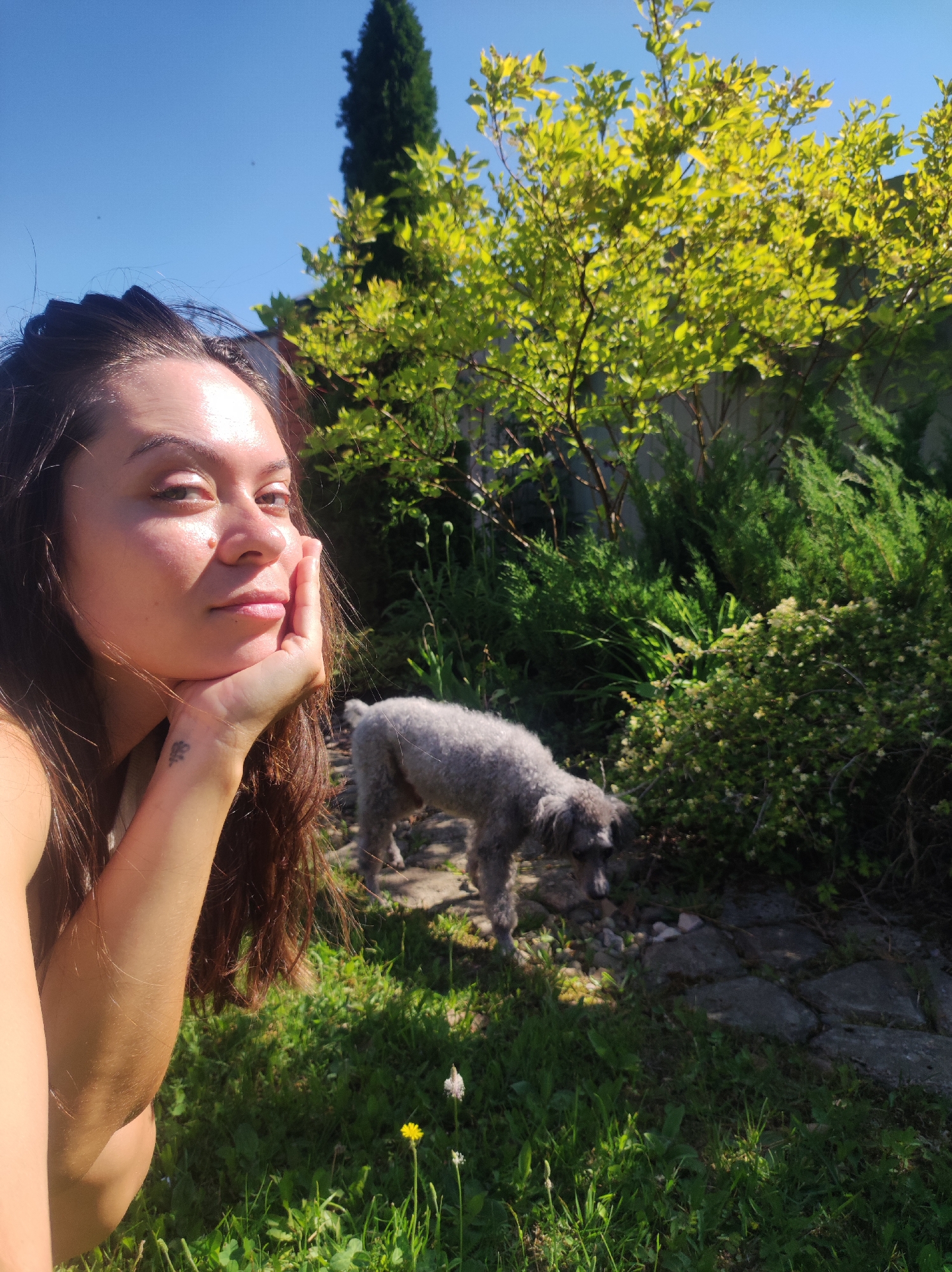 A young woman with long, dark hair is taking a selfie in a sunlit garden. She is resting her chin on her hand and looking towards the camera. Her left arm is visible in the foreground, and she has a small tattoo on her wrist. Behind her, a small, gray poodle stands on a stone path, looking down. The garden is lush with green grass, trees, and bushes, bathed in warm sunlight. The sky is a clear blue. The image appears to have been taken in a residential backyard, possibly in the city of Fokino, Russia, given the presence of mature trees and a well-maintained garden, common in such areas.