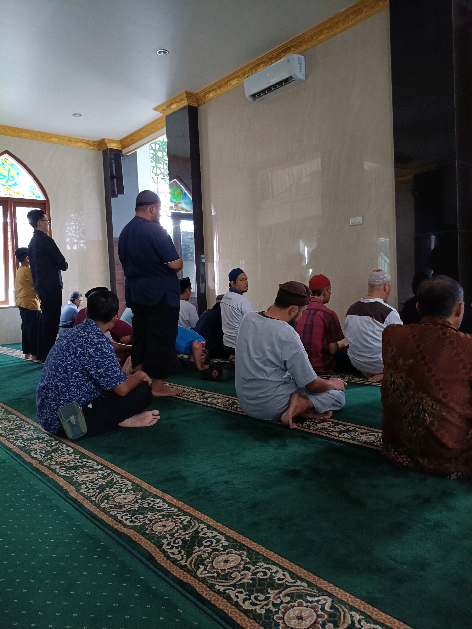 The image depicts a group of men engaged in prayer inside a mosque. The mosque has a green carpet with ornate borders. Several men are seated on the carpet, some in prayer positions, while others are standing. One man, wearing a blue patterned shirt and black pants, is seated facing away from the camera, with a small green pouch attached to his pants. To his left, another man in a gray shirt and black pants is also seated in prayer. In the background, more men are visible, some standing and some in prayer. The mosque's interior features light-colored walls, dark pillars, and decorative gold trim along the ceiling. An air conditioning unit is mounted on the wall. Stained-glass windows with geometric patterns are visible in the background, suggesting the location is the city of Maguwoharjo, Indonesia. The lighting is bright, indicating it is daytime.
