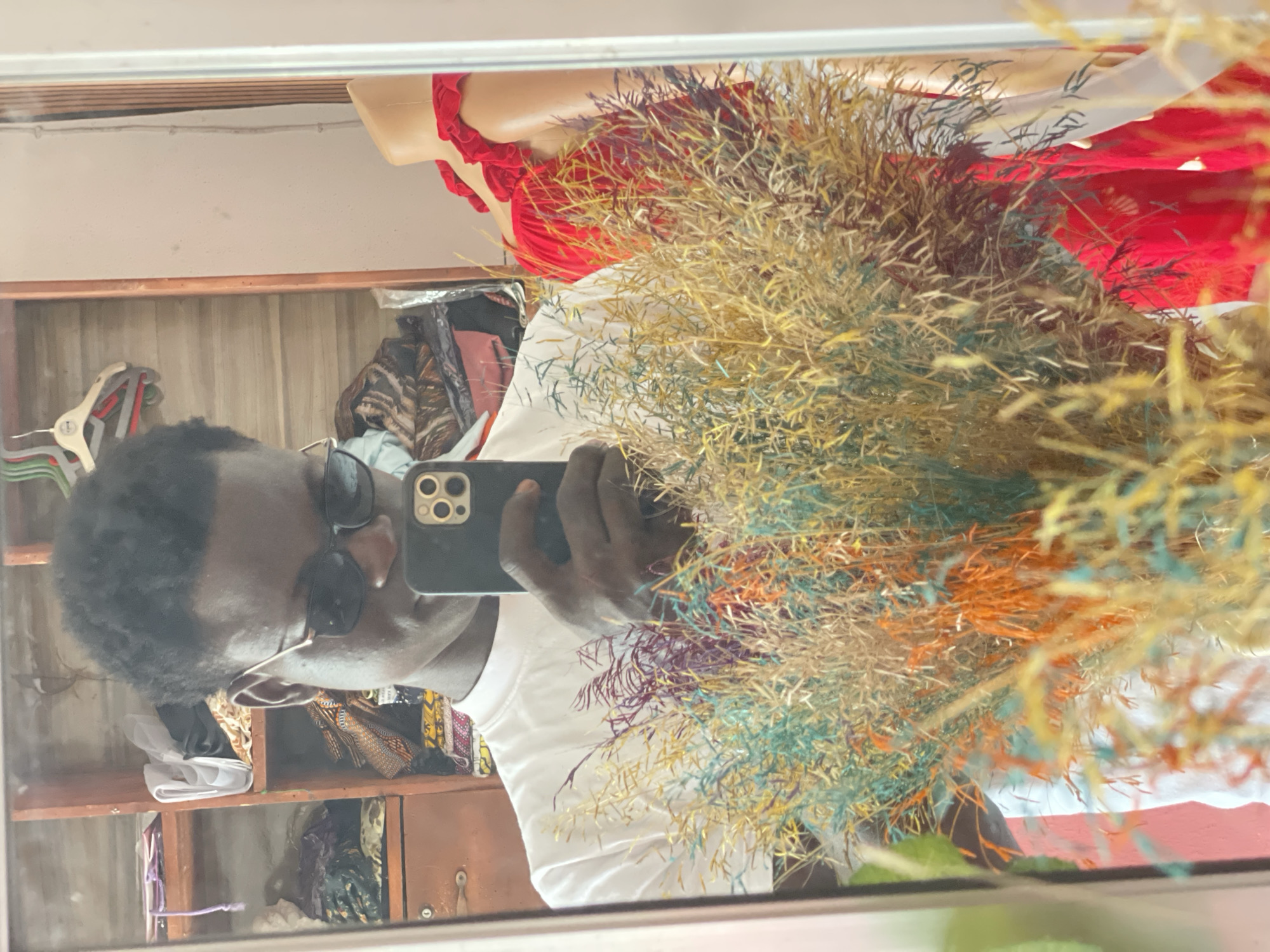 A person with dark skin and curly hair, wearing sunglasses and a white t-shirt, is holding a smartphone to take a selfie in a mirror. The background, visible through the mirror's reflection, includes a wooden shelf unit filled with clothing items, a mannequin torso with red garments, and a dried floral arrangement in the foreground. The reflection is set in what appears to be a shop in Badagry, Nigeria.