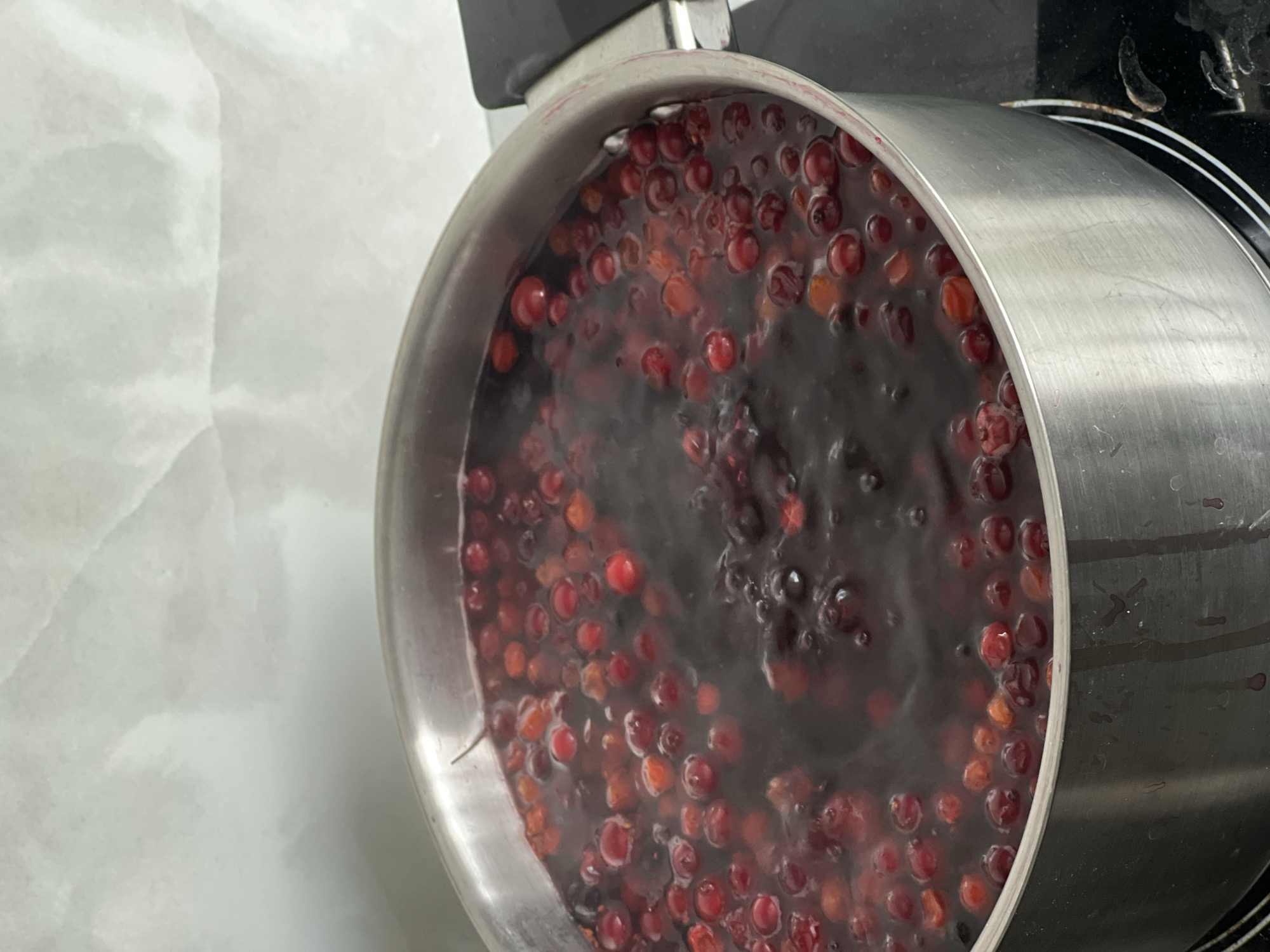 A stainless steel pot sits on a black stovetop. The pot contains a dark, simmering liquid with numerous red berries floating within. The background features a mottled, light-colored surface. It is impossible to determine a precise location, such as the city of Vologda, Russia, from the visual data provided.
