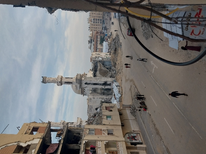 The image presents a wide view of a heavily damaged urban area in the city of Khan Yunis, Palestinian Territory, under a partly cloudy sky.
In the mid-ground, a prominent mosque with a light-colored dome and a tall, slender minaret stands. The mosque structure and its immediate surroundings exhibit significant damage, including visible structural compromise and rubble accumulation at its base. The top section of the minaret appears dark.
To the left of the mosque, a multi-story building is severely damaged, with large sections of its facade collapsed or missing, revealing interior spaces. Laundry is visible hanging on some balconies. The building's exterior shows multiple marks consistent with bullet impacts or shrapnel. Piles of debris are concentrated around the base of this structure. Further damaged buildings and extensive rubble characterize the background and the right side of the scene.
A two-lane paved road runs through the center, displaying faded white lane markings. In the foreground, a man in dark clothing walks on the road away from the viewer. Further down the road, on the left side, a group of approximately 5-7 individuals is gathered near the damaged building. Closer to the mosque, another group of 3-4 people is walking, with one person appearing to push a small cart. Two individuals are also seen riding bicycles on the road. Additional human figures are visible walking in the distance.
On the far right, partially obscured by prominent dark electrical or communication wires and cables, a person in a long dark red dress stands near a partially visible storefront sign featuring Arabic script, including "جراح" (Jarah). The areas flanking the road consist of disturbed earth and scattered debris.