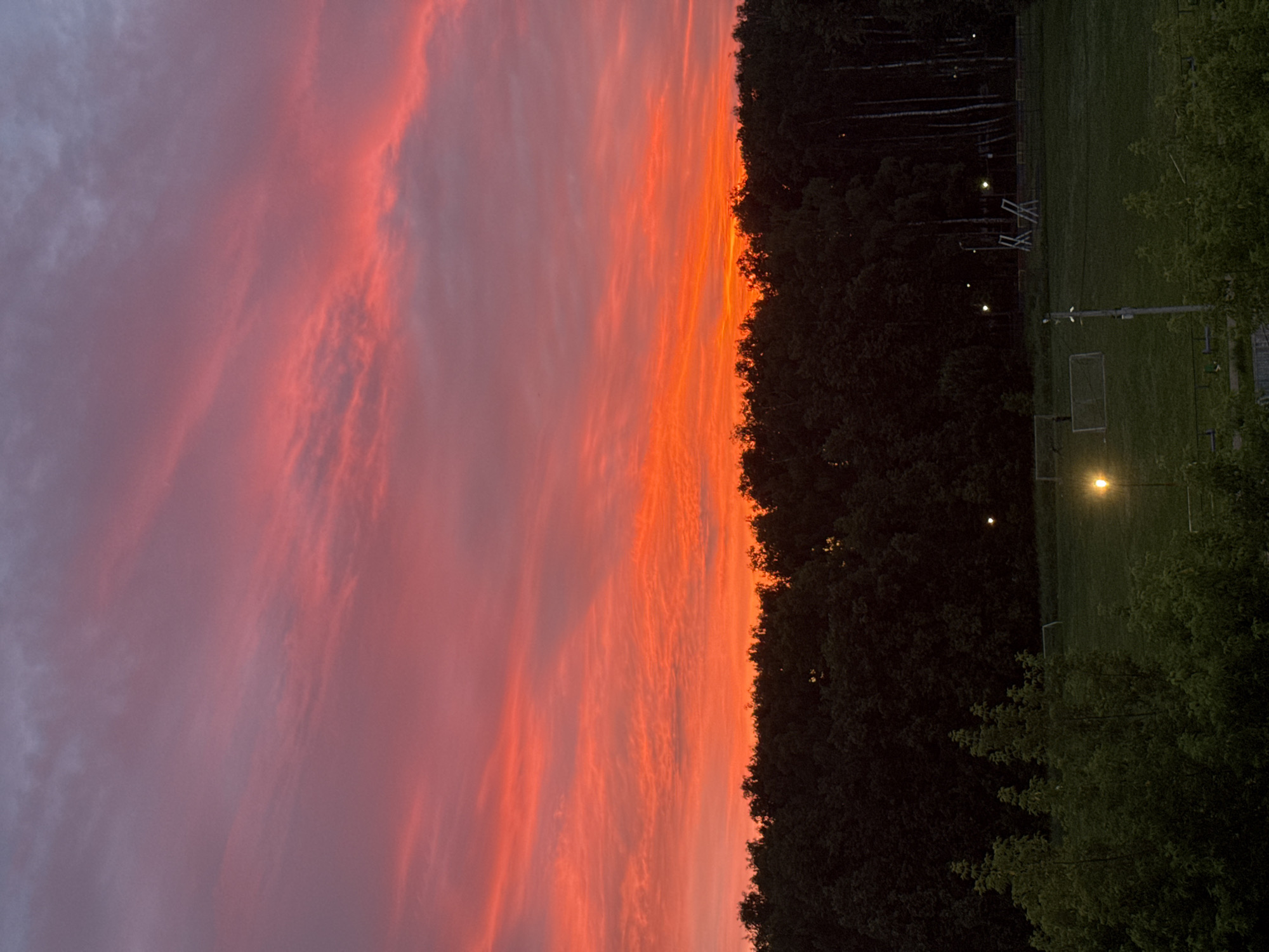 A vibrant sunset paints the sky with intense shades of orange, red, and pink, creating a dramatic backdrop over a dense line of dark green trees. Below the treeline, a grassy field is visible, featuring elements of a sports complex. A soccer goal with white netting is situated on the field, and several lampposts are scattered across the area, with two emitting a bright yellow light. The overall scene suggests an outdoor recreational space at dusk, with the natural beauty of the sunset contrasting with the man-made structures.