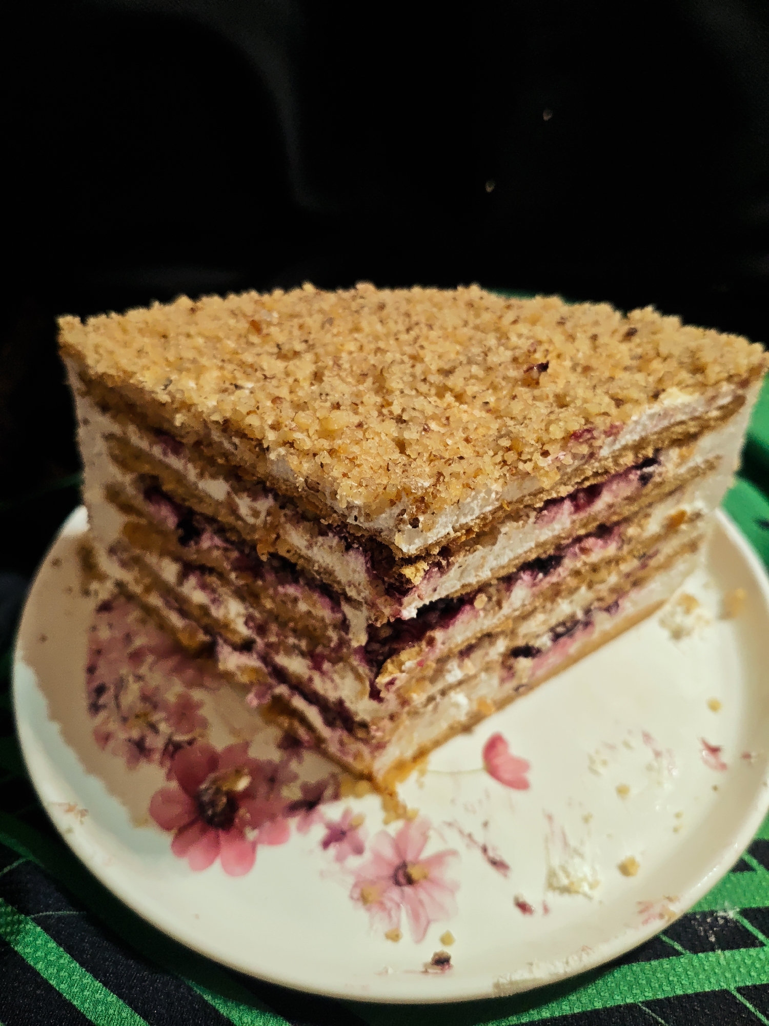 A triangular slice of layered cake sits on a white plate with pink floral designs. The cake consists of several layers of what appears to be a pastry or biscuit base, separated by layers of white cream and a dark-colored filling, potentially fruit or jam. The top layer is coated with a crumbly topping. The plate is resting on a dark-colored surface with green stripes. The background is a dark, blurry expanse. The city of Unknown, Unknown cannot be determined from the image.