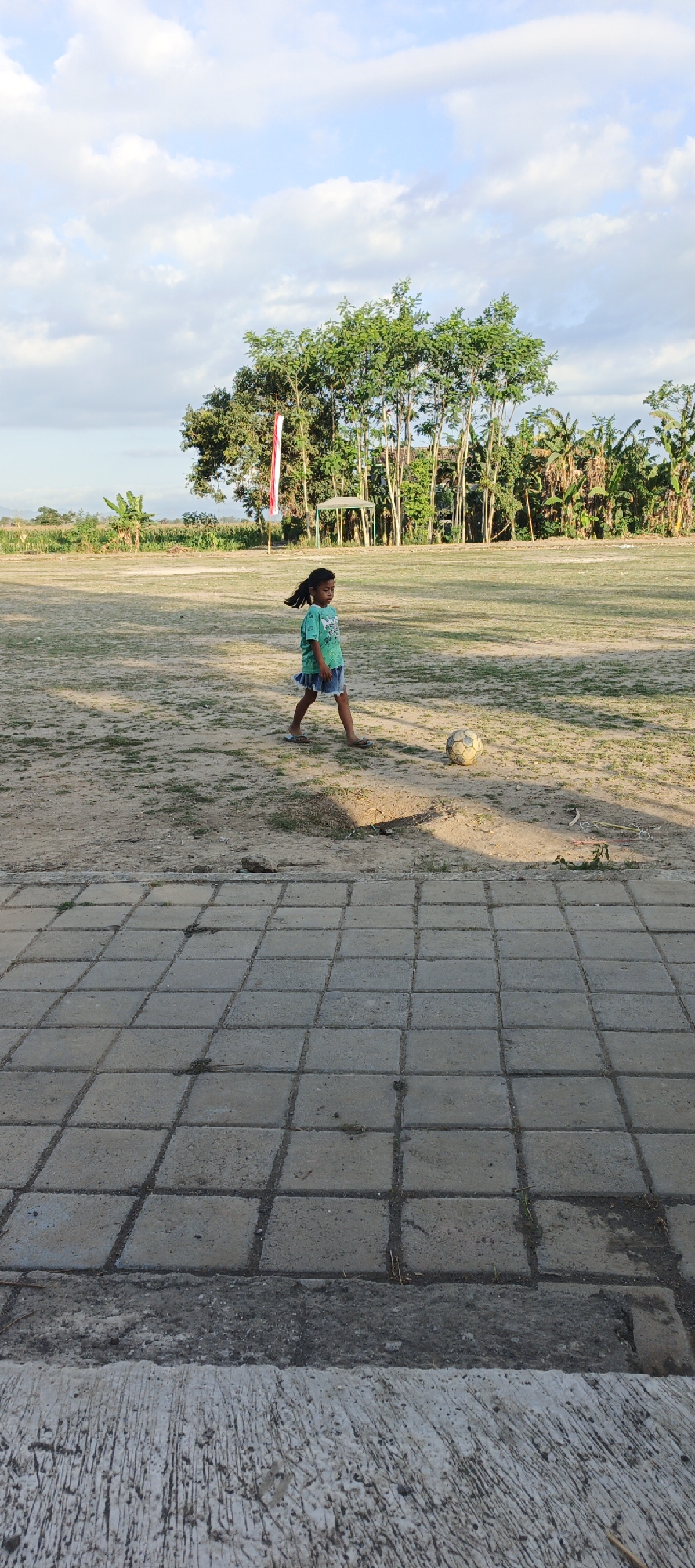 A young girl is playing with a soccer ball on a dirt field in Kasiyan Timur, Indonesia. The girl, wearing a green t-shirt and a blue skirt, is captured mid-stride, her long dark hair flowing behind her. The soccer ball, appearing weathered, rests on the ground to her right. The field is mostly bare dirt with sparse patches of grass and is bordered by trees in the background, including a tall flag pole with a red and white Indonesian flag. A hexagonal gazebo is visible behind the flag pole. The foreground is dominated by a paved area made of square concrete tiles, with a rough concrete border separating it from the dirt field. The sky above is a pale blue with scattered white clouds, and the lighting suggests it is either early morning or late afternoon.