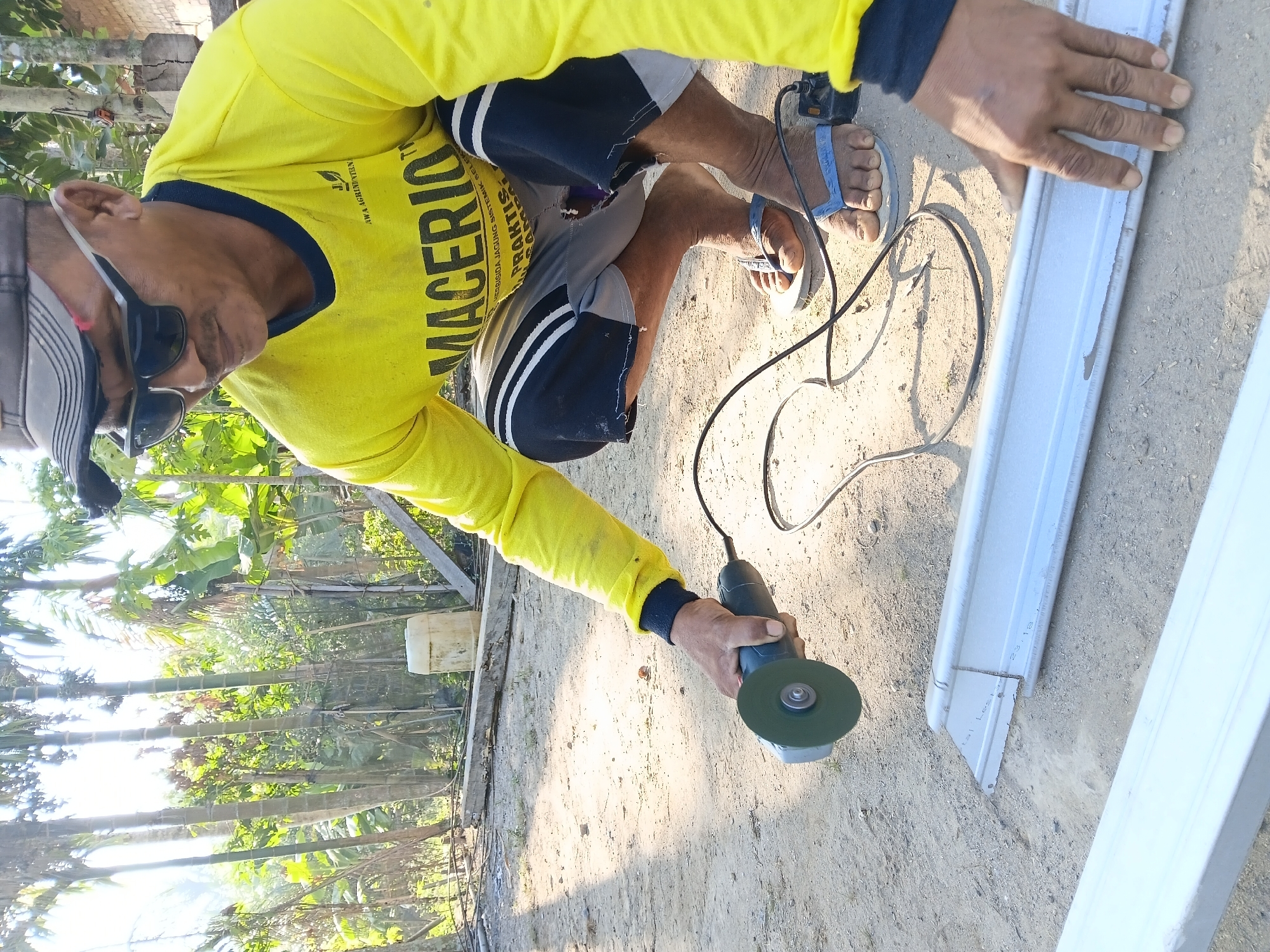 In Margo Rukun, Indonesia, a man wearing a yellow long-sleeved shirt and grey shorts squats on a dirt ground amidst a backdrop of palm trees and vegetation. He is using a green angle grinder to cut a section of a silver metal beam. A black cord from the grinder snakes across the ground. The man wears sunglasses and a grey cap. His feet are clad in blue flip-flops. The scene appears to be an outdoor worksite, likely in a rural or semi-rural setting given the natural surroundings.