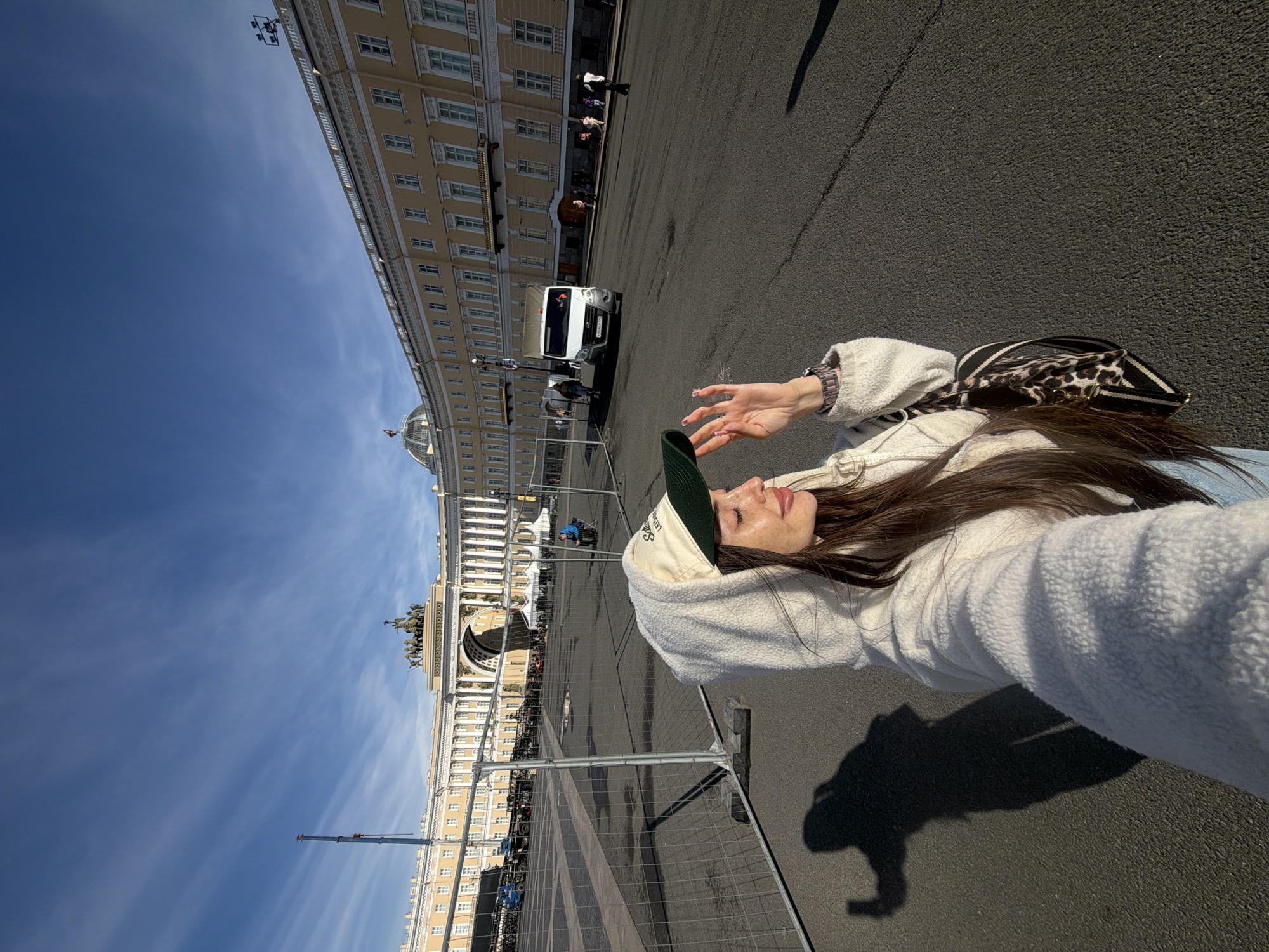 A medium shot captures a young woman standing outdoors in a public square. She is wearing a white hooded sweatshirt, a beige baseball cap with a green brim, and she has long, dark hair. Her right hand is raised, touching her face. A leopard-print bag with black and white straps is slung over her shoulder. Her shadow is cast on the asphalt ground to her left, holding what appears to be a phone, indicating she is taking a selfie.
In the background, a large, yellow, neoclassical building with numerous windows curves around the square. At its center is a large triumphal arch topped with a sculpture of chariots and horses. The building is partially fenced off with temporary metal barriers. Beyond the barriers, a white delivery van is visible, along with several pedestrians and a blue crane in the far distance. The sky above is clear blue with wispy white clouds.
The architectural style of the building and the triumphal arch strongly suggest the location is Palace Square in Saint Petersburg, Russia. The building is the General Staff Building, and the arch is the Triumphal Arch of the General Staff Building.