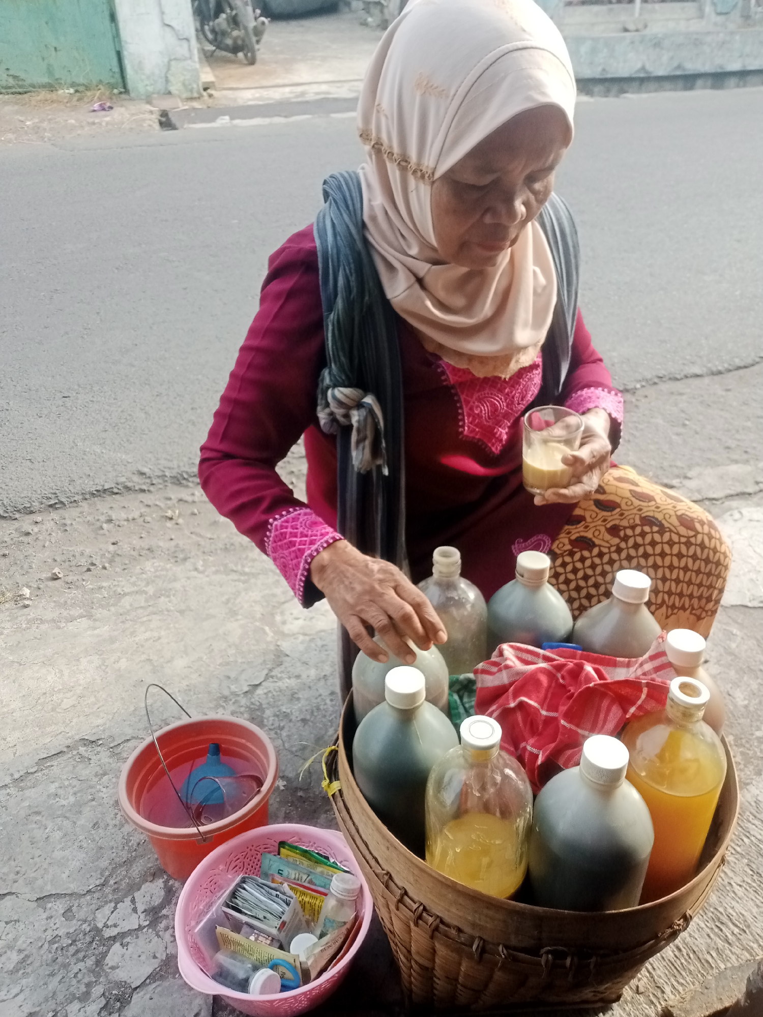 An elderly Indonesian woman wearing a light-colored hijab and a maroon long-sleeved top is seated outdoors, likely on a street in Tasikmalaya, Indonesia, as indicated by the presence of a motorbike and a typical Indonesian street scene. She is holding a glass of a light-colored beverage in her right hand and her left hand is reaching towards a basket filled with several large glass bottles. These bottles contain various liquids, some appearing to be yellow or amber in color, others dark. A red and white checkered cloth is placed in the basket. In front of the basket, there are two smaller containers: a red bucket with a blue funnel and a pink plastic basket filled with small packets and bottles, possibly containing spices or other small goods for sale. The ground is paved with concrete and asphalt. The woman's attire, the presence of goods for sale, and the street setting suggest she is a street vendor.
