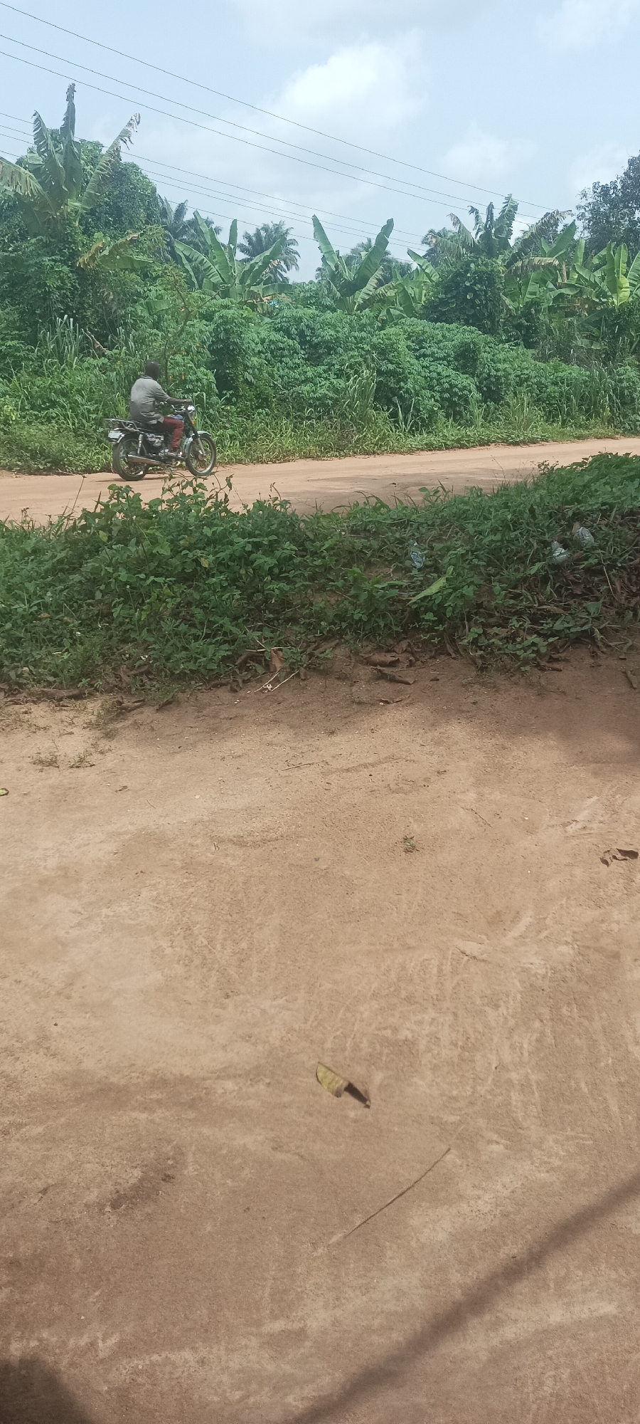 The image depicts a scene in Amauzu, Nigeria, along a dirt road lined with dense green vegetation. A man is riding a motorcycle on the road, moving from left to right. The foreground is dominated by a dirt area, possibly a yard or open space, with some scattered debris. Overhead power lines are visible in the sky.