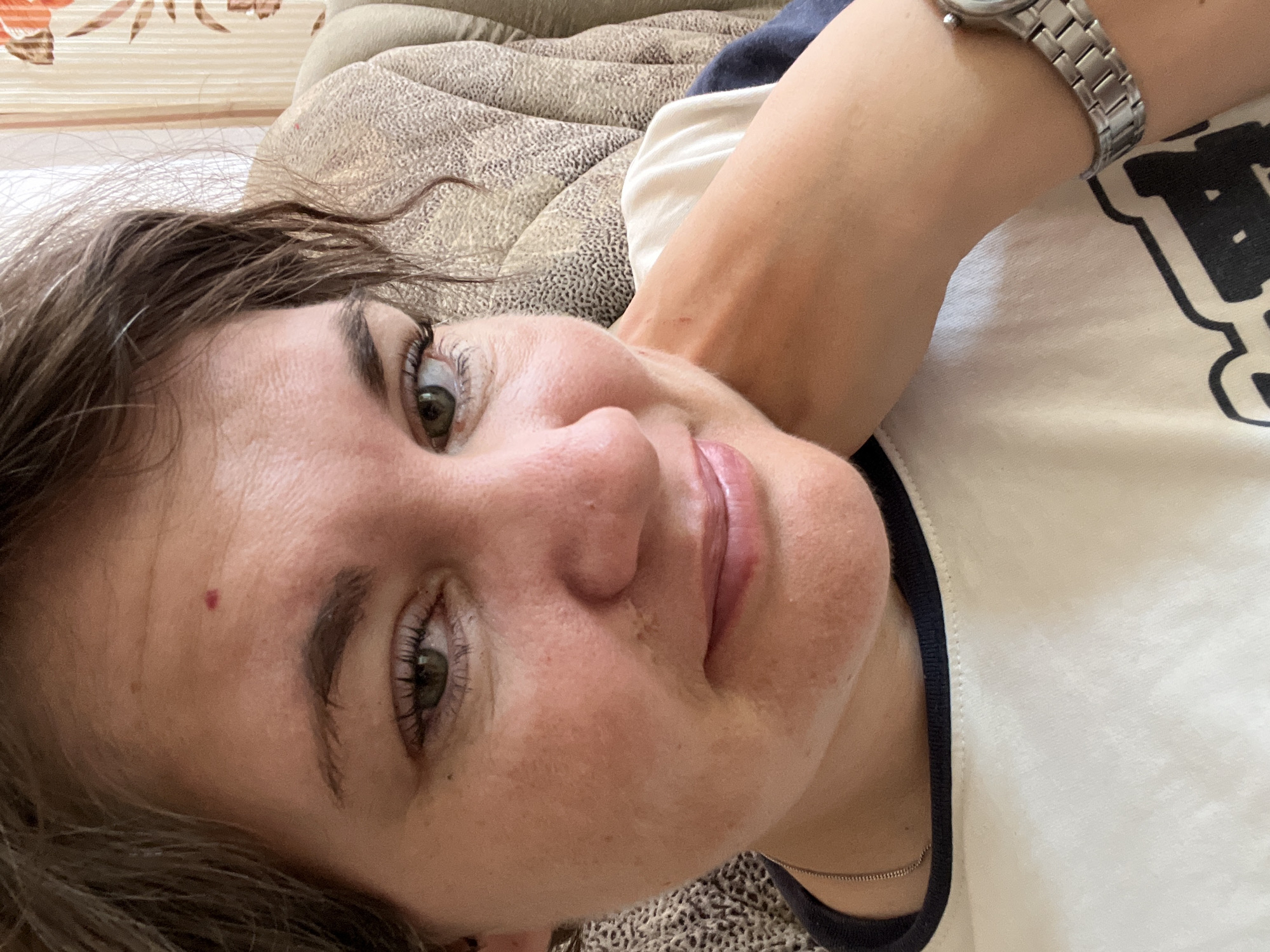 A close-up, slightly upward-angled selfie captures a woman from the chest up, in what appears to be an indoor setting, possibly a home in Afipskiy, Russia. The woman has short, dark brown hair, and her left hand is resting against her neck, supporting her head. She is wearing a light-colored t-shirt with a dark neckline and a silver watch on her right wrist. A thin silver chain is visible around her neck. The background includes a textured, upholstered piece of furniture in a neutral color and a curtain with a floral pattern featuring shades of orange and brown. The lighting suggests an indoor environment, possibly with natural light coming from a window. The overall impression is a casual, personal moment.