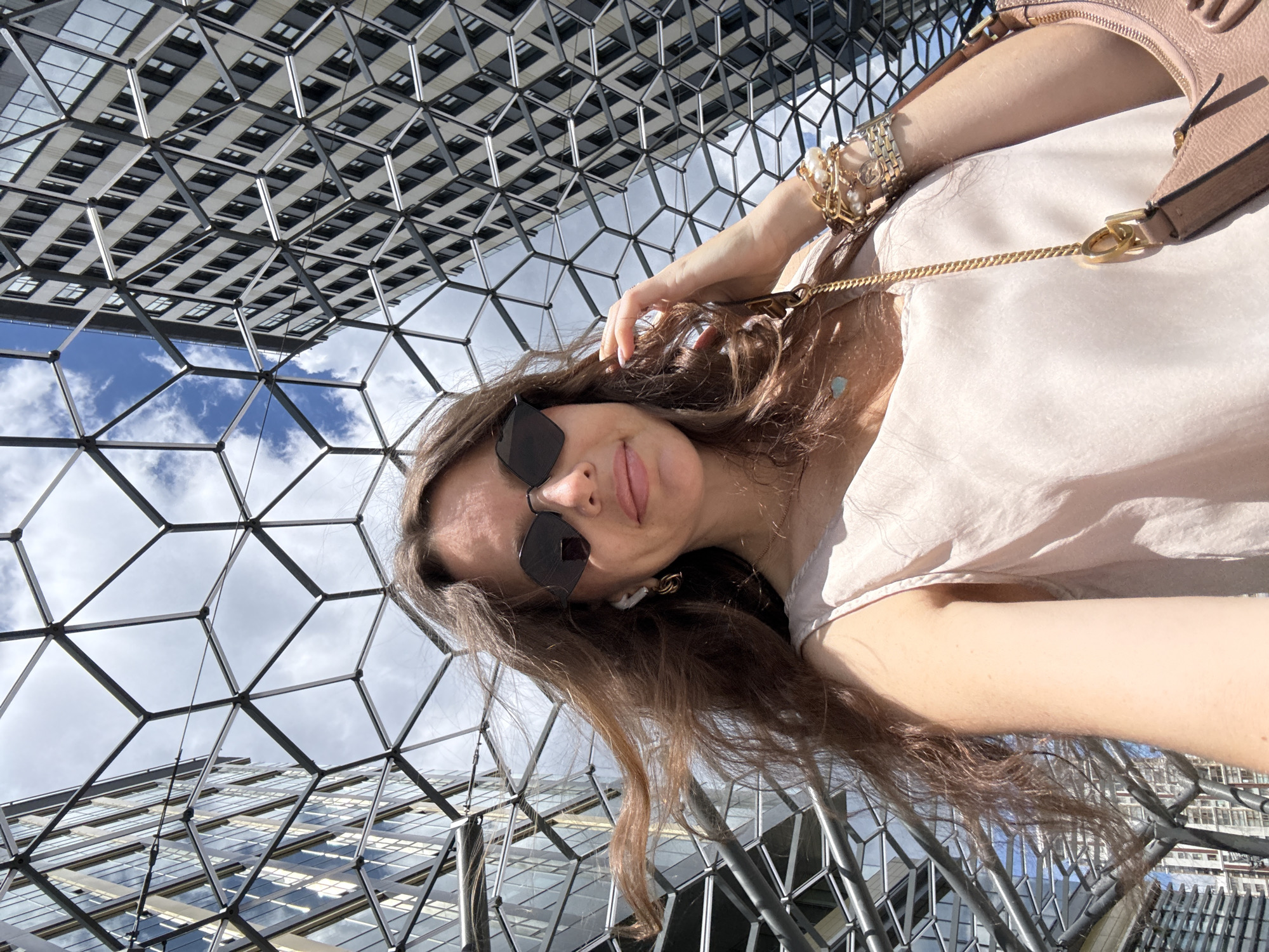 The media file is a selfie of a woman taken outdoors. She is positioned in the center of the frame, with her face turned towards the camera. She is wearing sunglasses and a light-colored top. A light-colored purse with a gold chain strap is visible on her shoulder. Her left arm is adorned with several bracelets, including a watch and chain-link bracelets with pearl embellishments.
The background is dominated by a striking modern architectural structure with a geometric pattern of intersecting metal beams and glass panels, reflecting the sky and clouds. The sky appears bright with scattered clouds, suggesting it is daytime. The woman's expression is relaxed and her pose suggests she is casually taking a photo of herself. The overall impression is one of a bright, sunny day in a contemporary urban environment. There is no visible text in the media file. The location context is Moscow, Russia.