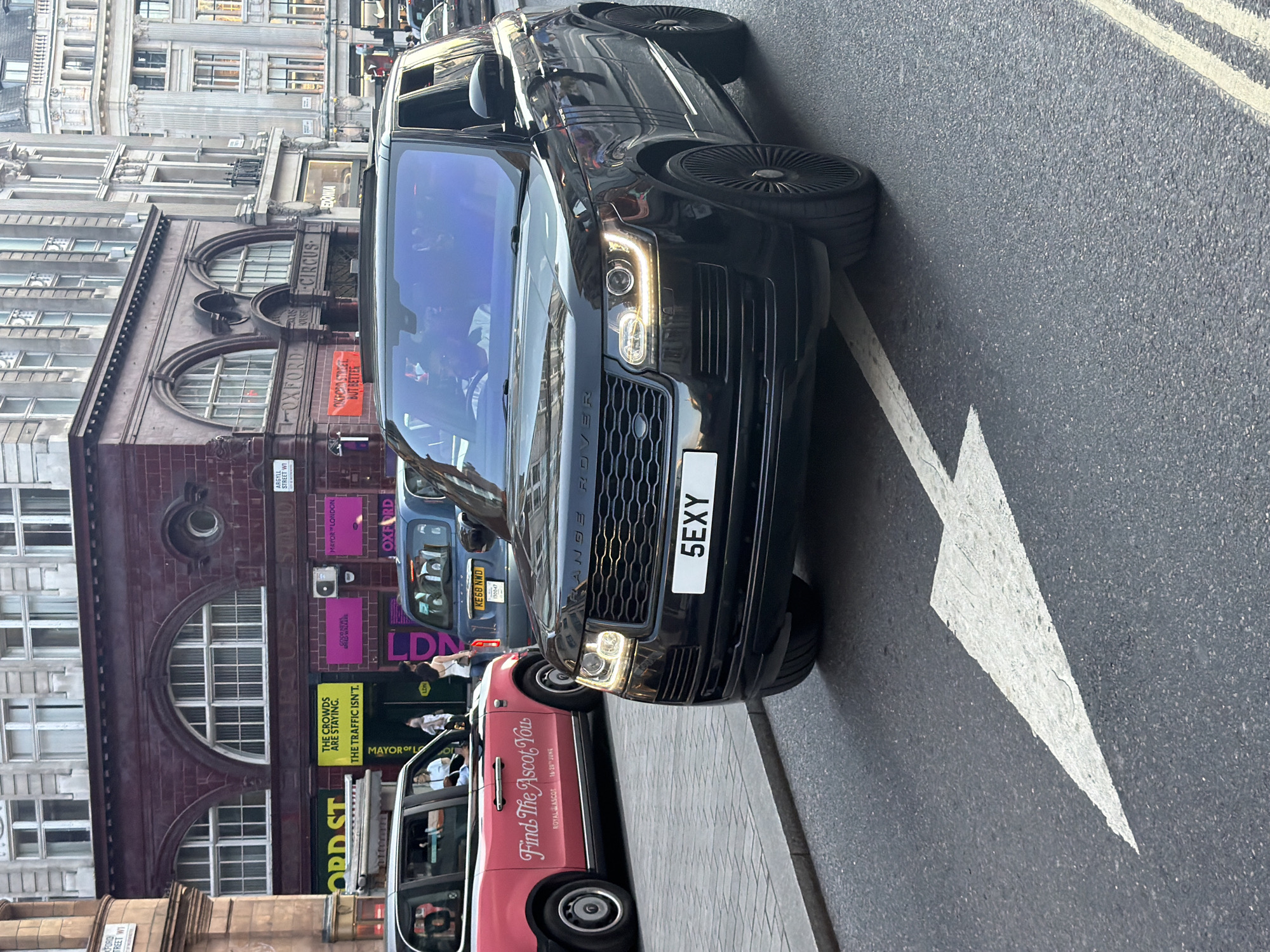 This is a street-level, eye-level, medium shot of a black Range Rover driving on Oxford Street in London, United Kingdom. The car has the license plate "5EXY." A pink taxi with "Find The Ascot You" written on its side is to the left of the Range Rover. In the background are buildings with arched windows and signs for "Oxford Circus" and "Oxford Street." The day appears to be overcast.