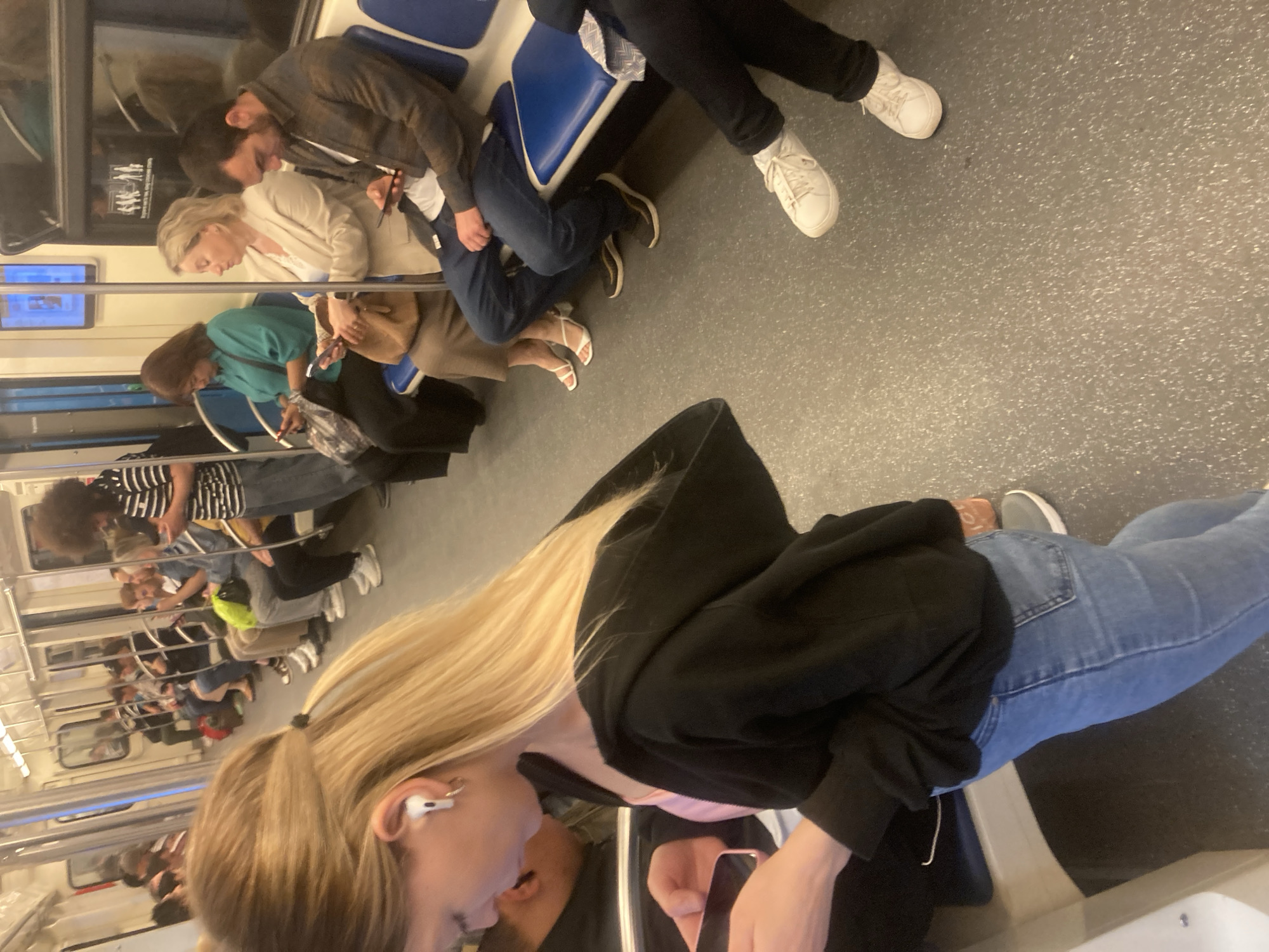The image captures the interior of a metro train carriage, likely in Moscow, Russia, identifiable by the design of the train cars and the general public transportation system. Several passengers are visible, both seated and standing, engaging in typical activities such as using mobile phones, and appearing to be commuting. The lighting is somewhat diffused, typical of underground transit systems. The train car appears to be in motion. The foreground highlights a young woman with long blond hair, wearing an earbud and holding a mobile phone, facing towards another person, possibly engaged in a conversation. The scene depicts an everyday occurrence within an urban transit setting.