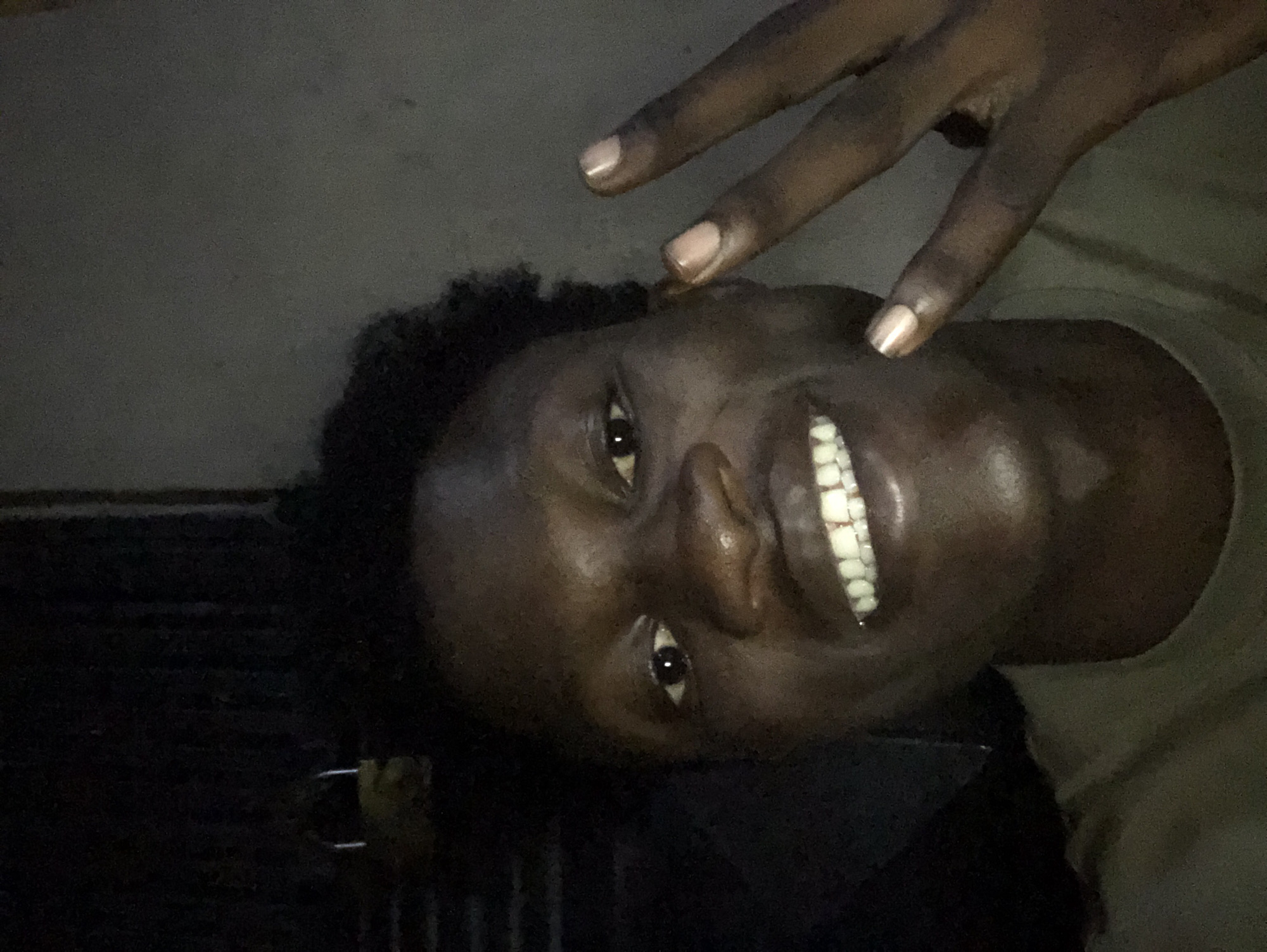 A close-up, head-and-shoulders selfie shows a young Black male with dark skin and short, dark, curly hair. He is smiling broadly, revealing white teeth, some of which appear to have braces. His right hand is raised, with two fingers extended in a peace sign, and the thumb tucked down. The background is dark and indistinct, with some vertical lines visible on the left and a lighter, textured surface on the right. A yellow padlock is partially visible on the far left, hanging from something dark and vertical, possibly a door or gate. The lighting is dim, creating shadows on the subject's face and the background. The image appears to have been taken indoors or in a sheltered outdoor space at night or in low-light conditions. Given the context of Ado, Nigeria, this could be a residential area or a commercial establishment.