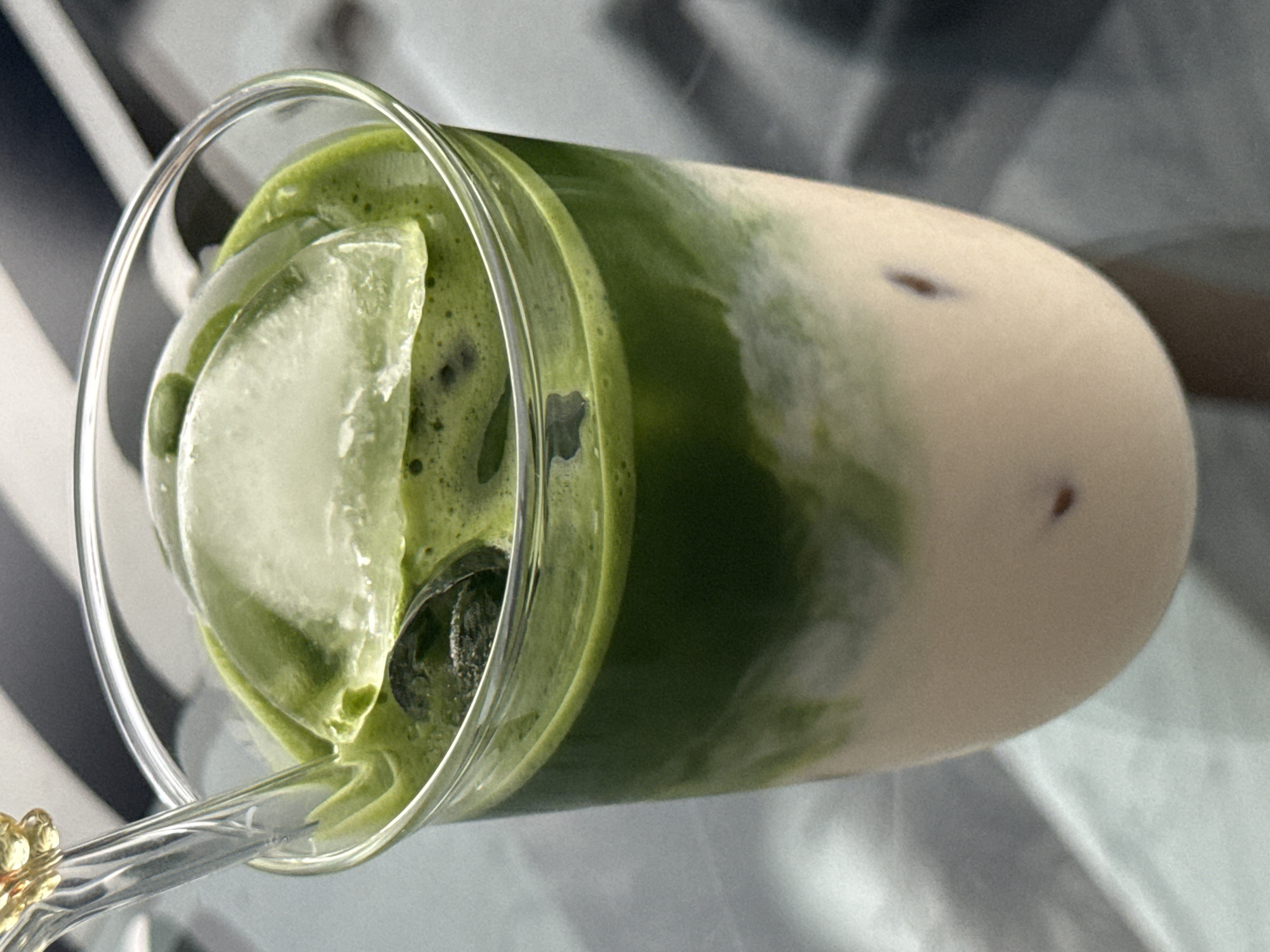 The image presents a close-up, horizontal view of an iced beverage served in a clear glass. The drink features two distinct layers: a vibrant green layer, likely matcha, occupies the top and left side of the glass, while a creamy, off-white layer, indicative of milk, fills the bottom and right. The layers show natural marbling and swirls where they meet.
Several large, clear ice cubes are submerged within the green matcha layer, some appearing frosty. Small, dark specks are visible throughout the green portion of the drink, and a few are also present in the white, milky section. A transparent straw or long spoon, featuring a decorative, possibly gold-toned, handle, is visible at the bottom left, extending into the glass. The background is softly blurred, displaying abstract patterns of light and dark, possibly originating from a striped surface or textile. The overall lighting is bright and even, highlighting the fresh appearance of the beverage. There is no visible text in the image.