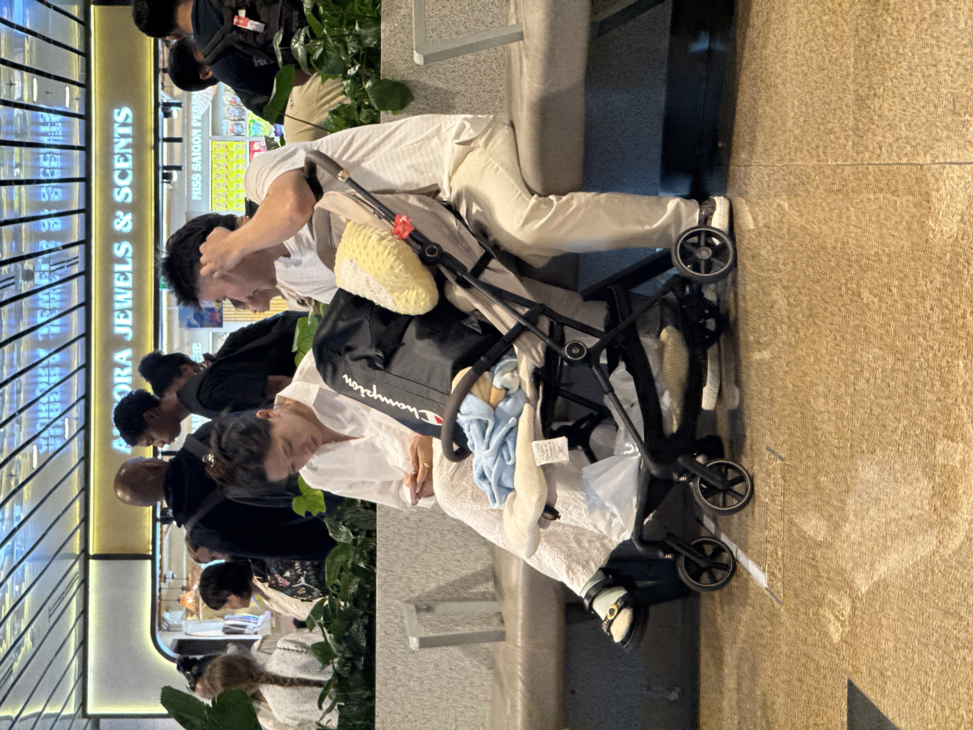 At the Cam Ranh International Airport, a couple sits on a gray bench in a brightly lit terminal. The woman, wearing a white dress and sandals, holds a smartphone and looks down at it. Beside her, a man in a white t-shirt and beige pants reaches for his ear. A stroller, carrying a yellow pillow and blue blanket, is positioned between them. In the background, a sign reads "AURORA JEWELS & SCENTS" and other shop signs. Plants are visible along the left side of the image.