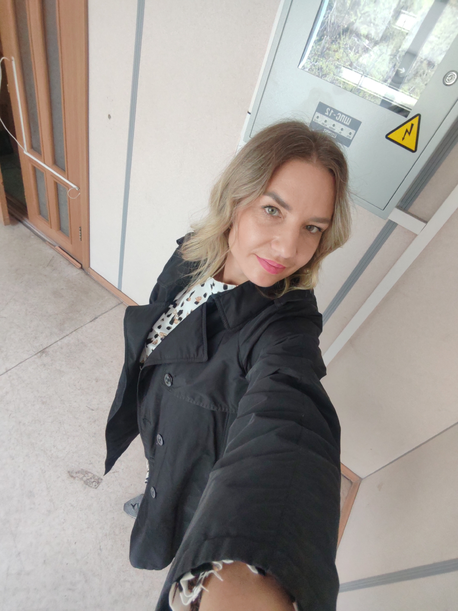 A woman is posing indoors, looking at the camera. She is wearing a black jacket over a patterned top. Behind her is a door, walls, and a grey electrical box with a yellow lightning bolt symbol. The location in the city of Unknown, Unknown cannot be determined.