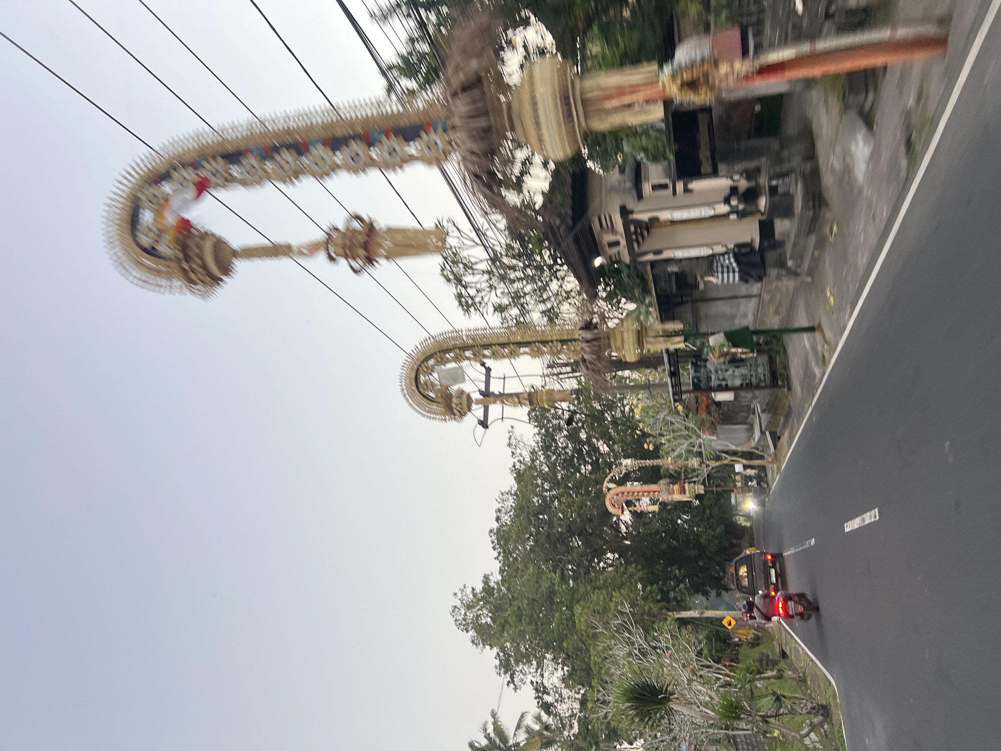This is a daytime, eye-level, full-shot image taken from a moving vehicle on a road in Tembuku, Indonesia. The scene captures a street lined with traditional Balinese offerings, known as Penjor, which are tall, curved bamboo poles adorned with woven palm leaves and decorations. Several Penjor are visible along the right side of the road, some with intricate detailing and colorful elements.
The road itself is paved and has white lane markings. On the left side of the road, there are trees and vegetation, including palm trees. A few buildings are visible behind the Penjor, suggesting a residential or commercial area.
Traffic is present in the image. A motorcycle is seen in the foreground, moving away from the viewer, with its taillights illuminated. Further down the road, a black pickup truck is also visible, as well as a motorcycle parked on the right side. The sky is overcast with a light blue hue. The overall impression is of a street in a tropical village during the day, decorated for a cultural or religious event.