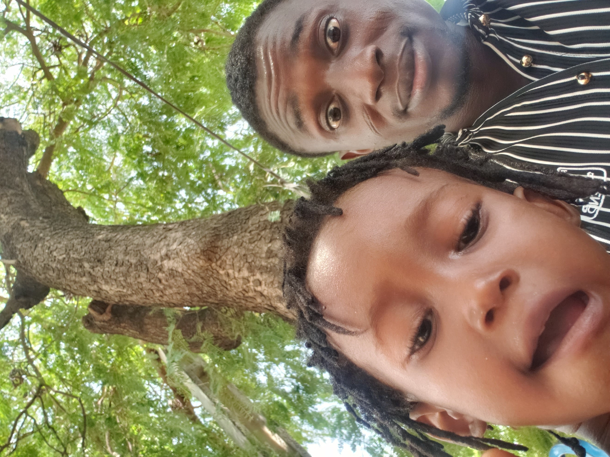 This image is a close-up, upward-angled shot of an adult male and a child, presumably taken as a selfie outdoors in Adankolo, Nigeria. Both subjects are positioned sideways in the frame, with the adult male dominating the upper portion and the child in the lower half.
The adult male, with short dark hair, looks directly at the camera with wide, expressive eyes and an open mouth, conveying a sense of surprise or playful enthusiasm. He wears a black and white striped shirt with prominent gold buttons on the placket. Below him, the child's face is partially visible, also looking towards the camera with an open mouth and dark dreadlocks. Their expression appears curious or engaged in the playful interaction.
The background features the bright green foliage of a tree canopy and a substantial tree trunk, indicating an outdoor setting, likely under the shade of a large tree. The bright illumination suggests it is daytime. The image captures a candid and affectionate moment between the two individuals. There is no visible text in the image.