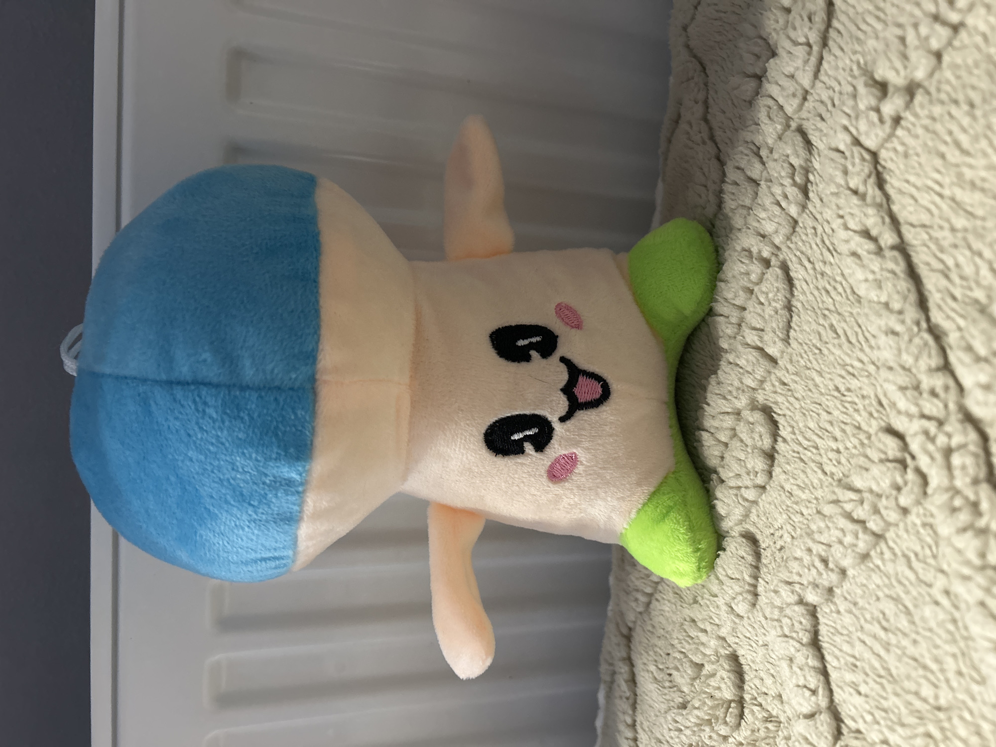 A plush toy, resembling a mushroom with a blue cap, peach-colored body, and green feet, sits on a textured blanket. The toy has embroidered black eyes, a smiling mouth, and pink blush marks on its cheeks. Behind the toy, a white radiator and a dark grey wall are visible. The scene is indoors and does not provide any specific indicators of being located in the city of Perm, Russia.