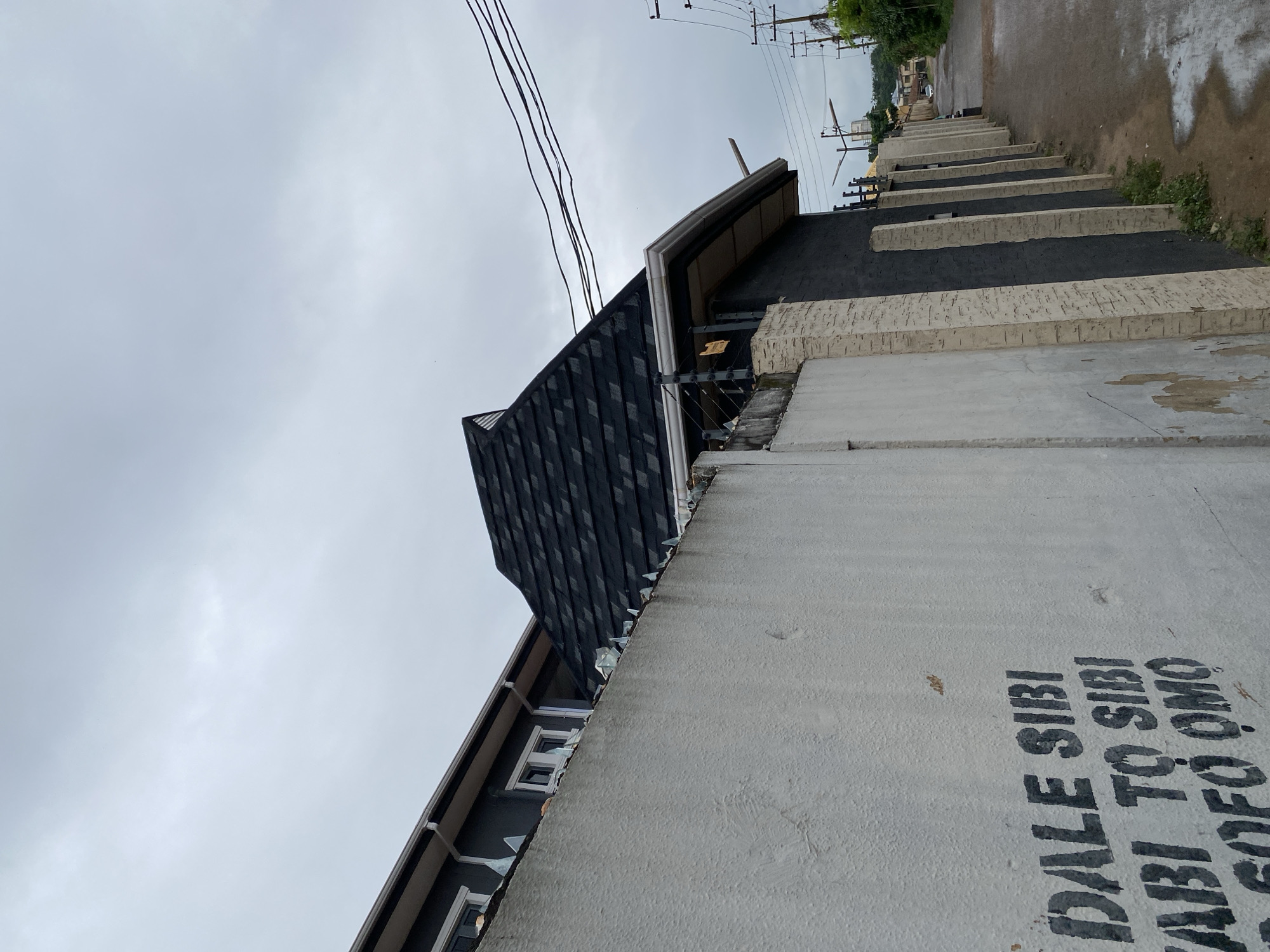The image depicts a residential property in Abeokuta, Nigeria, on a cloudy day. The foreground is dominated by a white, textured wall with black spray-painted text that appears to be in Yoruba. The text reads "DALE SIBI", "ABI TO SIBI", and "SOFO OMO".
Behind the wall, a dark gray building with a dark shingled roof is visible. The roof has a steep, pyramid-like section. Along the top of the white wall, shards of glass are embedded, indicating a security measure. The building has white trim around the roofline and several windows.
To the right of the building, a paved road is wet, suggesting recent rain. A line of utility poles with power lines runs along the side of the road. In the distance, more buildings and greenery are visible. The overall impression is of a secured residential area. There are no people visible in the image.