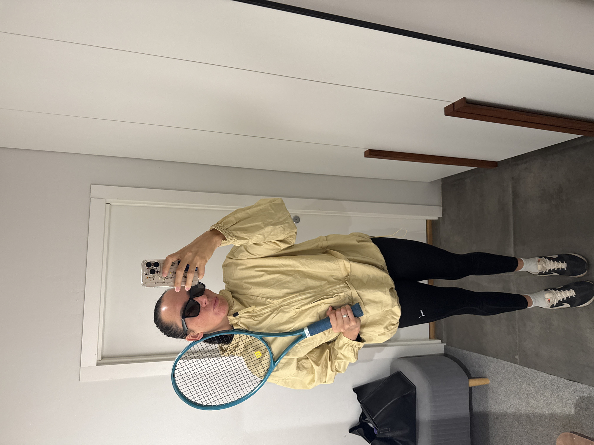 A woman stands in a room, holding a tennis racket and her phone, taking a mirror selfie. She is wearing a tan hooded jacket, black leggings with a Puma logo, white socks, and black and white sneakers. She has on sunglasses and her hair is pulled back in a ponytail. A black bag is visible on a grey ottoman to her left. The room appears to be a hallway or dressing area, with a white door and a mirrored wardrobe in the background. The floor is tiled.