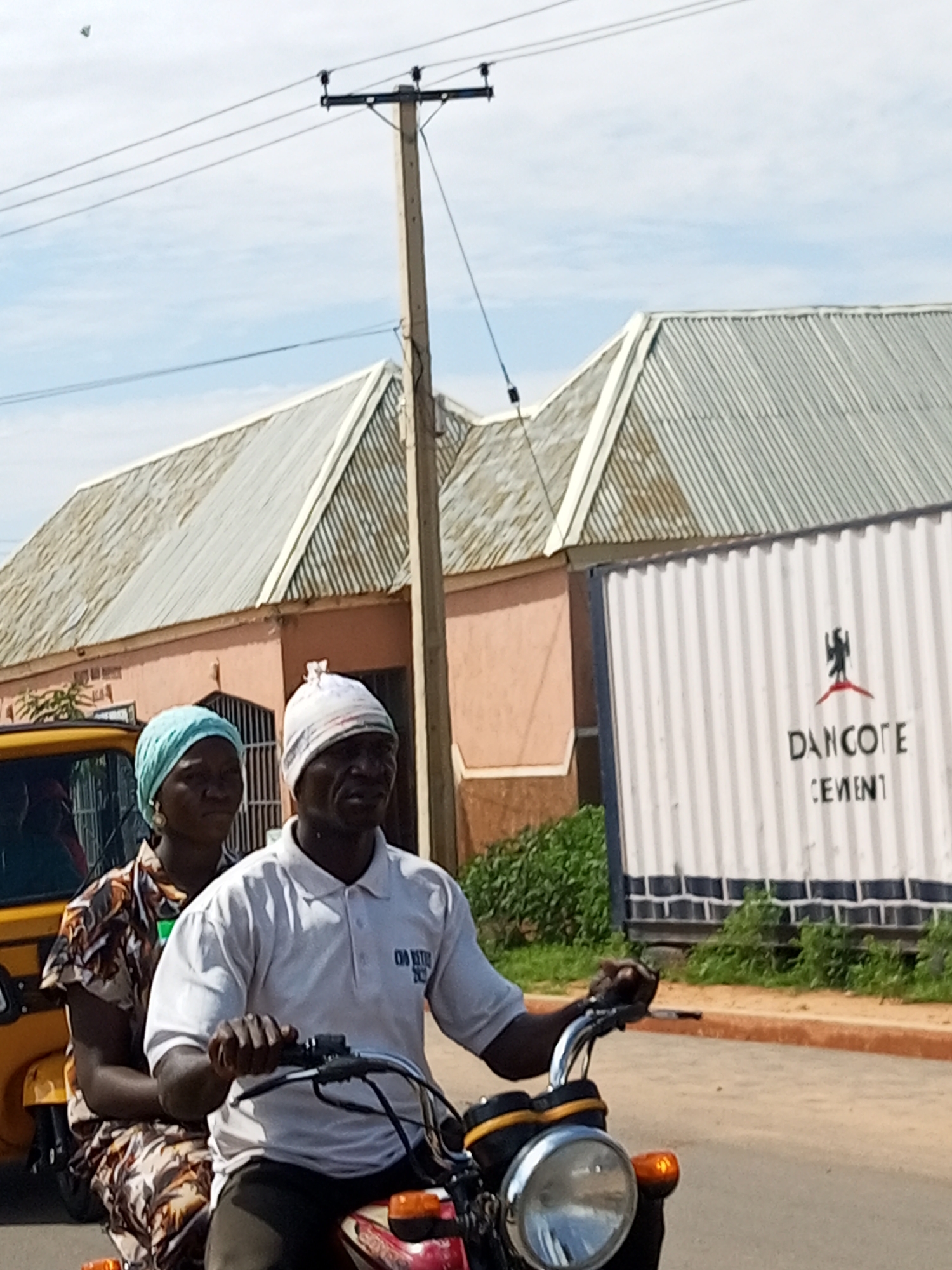 Two individuals, a man and a woman, are riding on a motorcycle on a paved road in Tempure, Nigeria. The man is driving the motorcycle, and the woman is seated behind him. The man is wearing a white polo shirt and a white cap, while the woman is wearing a patterned dress and a blue headscarf. In the background, there is a yellow tricycle, a pink building with a corrugated metal roof, and a white shipping container with "DANCOTE CEMENT" written on it. A utility pole with power lines stands to the left of the pink building. Green foliage is visible on the right side of the image. The sky is cloudy.