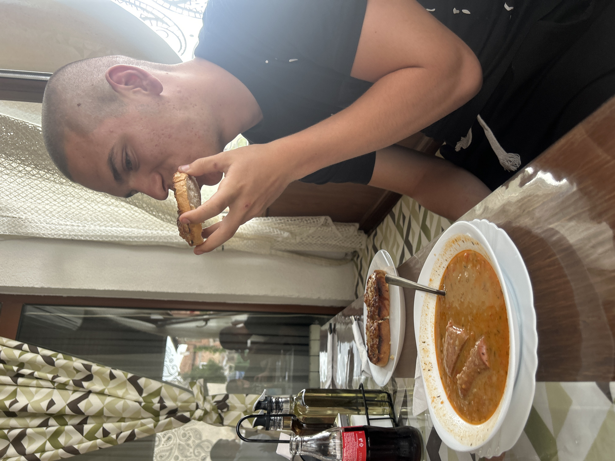 This image captures an indoor dining scene in Bania, Bulgaria, depicting a young man in the midst of enjoying a meal. The setting appears to be a restaurant or a home dining area, illuminated by bright daylight streaming through a large window.
The main subject is a male with a shaved head, wearing a black t-shirt, focused on eating. He is holding a piece of crusty bread to his mouth, about to take a bite. The meal laid out on the glossy wooden table includes a white bowl filled with a rich, reddish-brown stew or thick soup containing visible chunks of meat and possibly vegetables, with a spoon resting in it. Alongside the bowl, an oval white plate holds two halves of a baked bread roll, which appears to be the type the man is consuming.
Beverages on the table include a classic glass bottle of Coca-Cola, identifiable by its red label and logo. Next to it, a metal rack holds two other bottles, likely oil (clear liquid) and vinegar (dark liquid), commonly used as condiments. The table is partially covered by a patterned tablecloth featuring geometric shapes in shades of green, brown, and off-white. Matching patterned curtains are tied back to the side of the window, revealing a blurred view of what appears to be residential buildings or houses outside, consistent with a town or village environment. The scene portrays a casual moment of dining and nourishment.