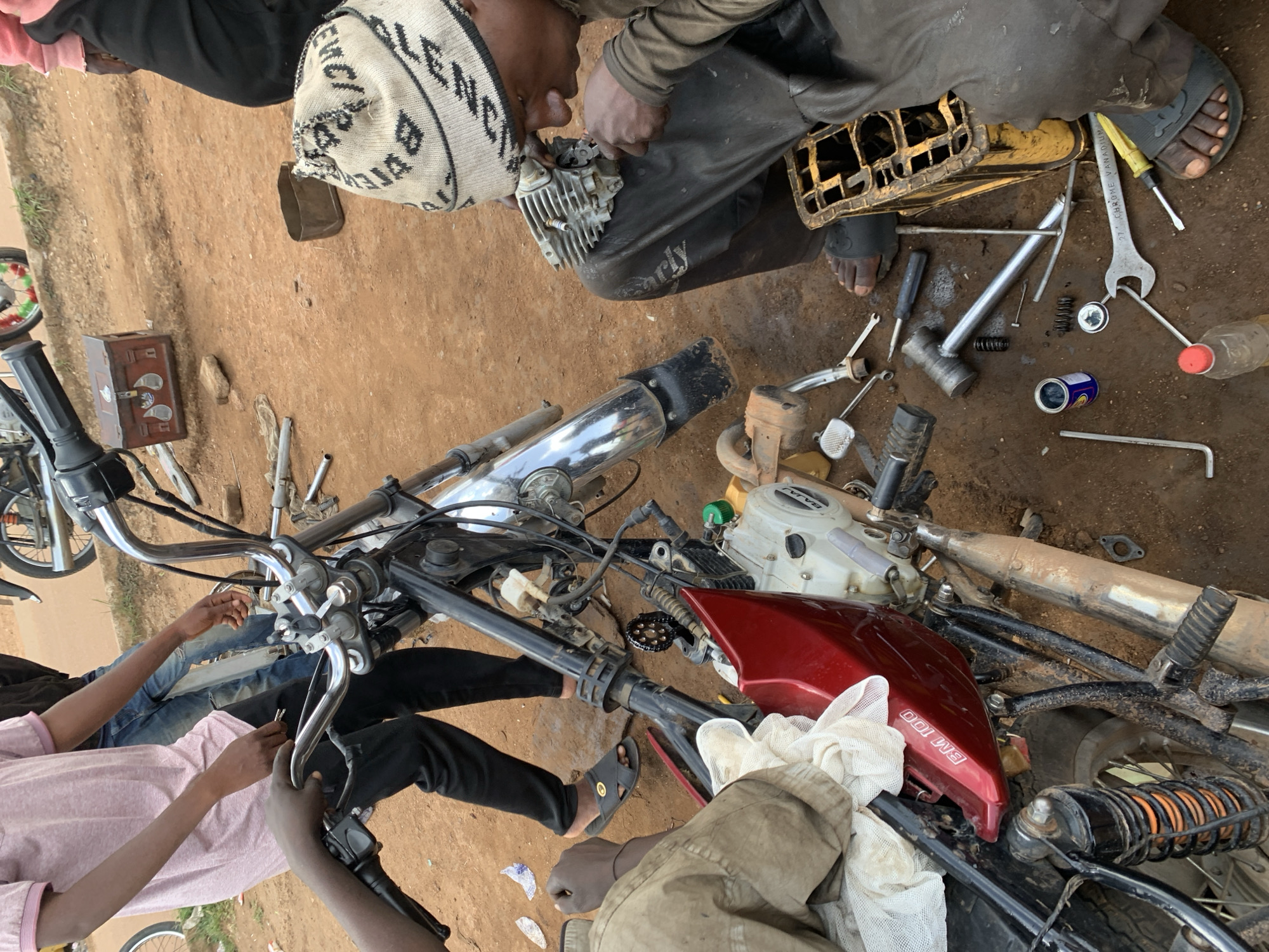 In Barakin Lerawi, Nigeria, a scene of motorcycle repair unfolds on a dirt ground. A person wearing a pink t-shirt and black pants stands next to a motorcycle, their hands engaged with the handlebars. Another individual, seated and wearing a patterned beanie and dark clothing, works on a motorcycle engine part. Scattered around them are various tools, including wrenches, a hammer, screwdrivers, and a can of oil, suggesting a workshop setting. The motorcycle itself appears partially disassembled, with its red fuel tank prominent. The ground is a mix of reddish-brown dirt and debris.
