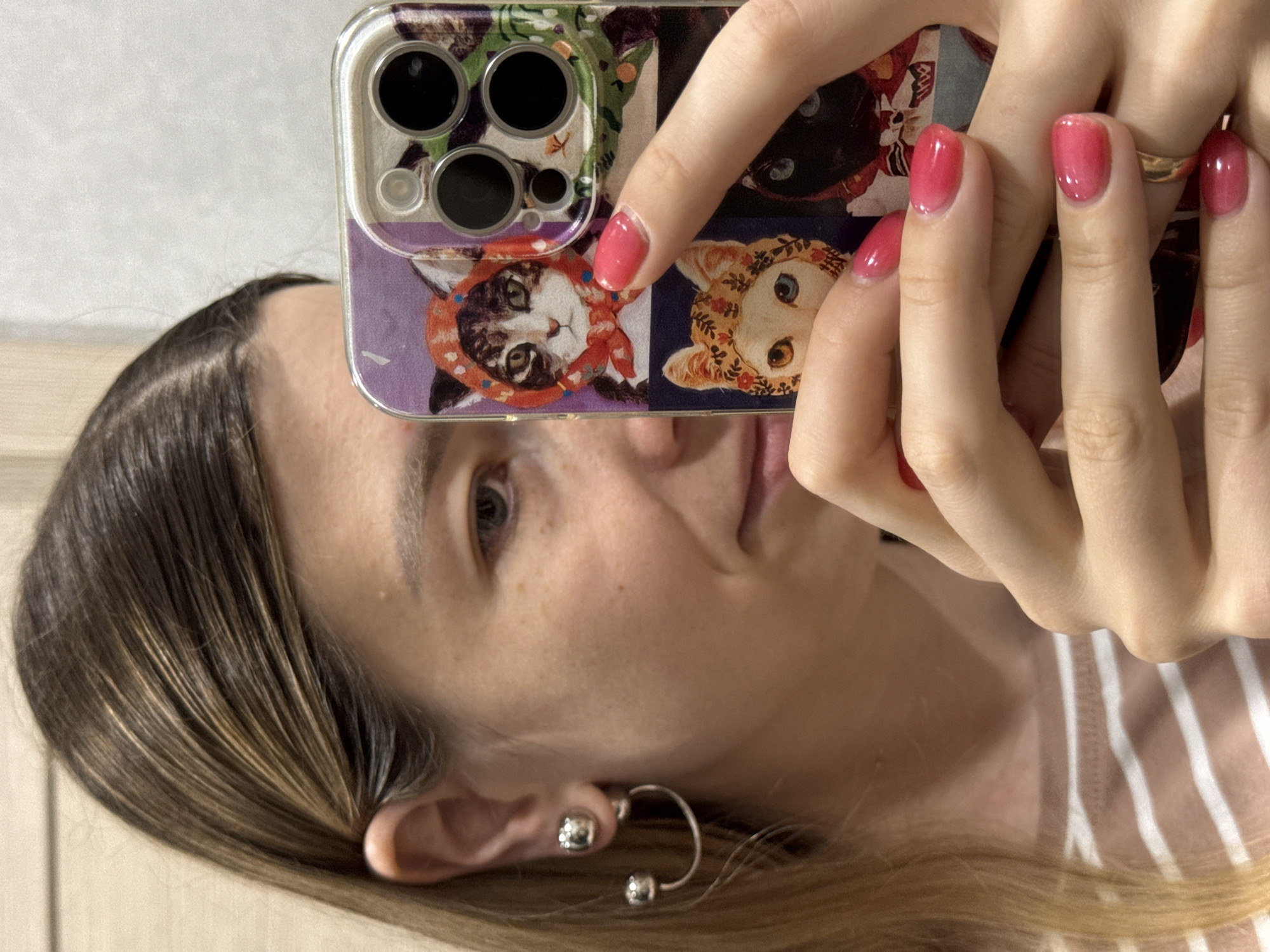 The image shows a close-up, upside-down selfie taken by a person. The person is holding a smartphone with a colorful case featuring illustrations of cats. Their hands, with freshly manicured pink nails and a gold ring, are visible as they hold the phone. The phone's camera array is also visible in the upper part of the image.
The person's face is partially visible, with one eye and a portion of their mouth showing. They are wearing a silver hoop earring with a small ball accent and a smaller stud earring on their earlobe. Their hair, which appears to be brown with blonde highlights, is also visible, framing their face. The background is blurred but suggests an indoor setting, possibly a room with a wall and a piece of furniture. The person is wearing a striped top. The overall mood appears casual and personal, typical of a selfie. There is no visible text in the image. The lighting suggests it was taken during the daytime.