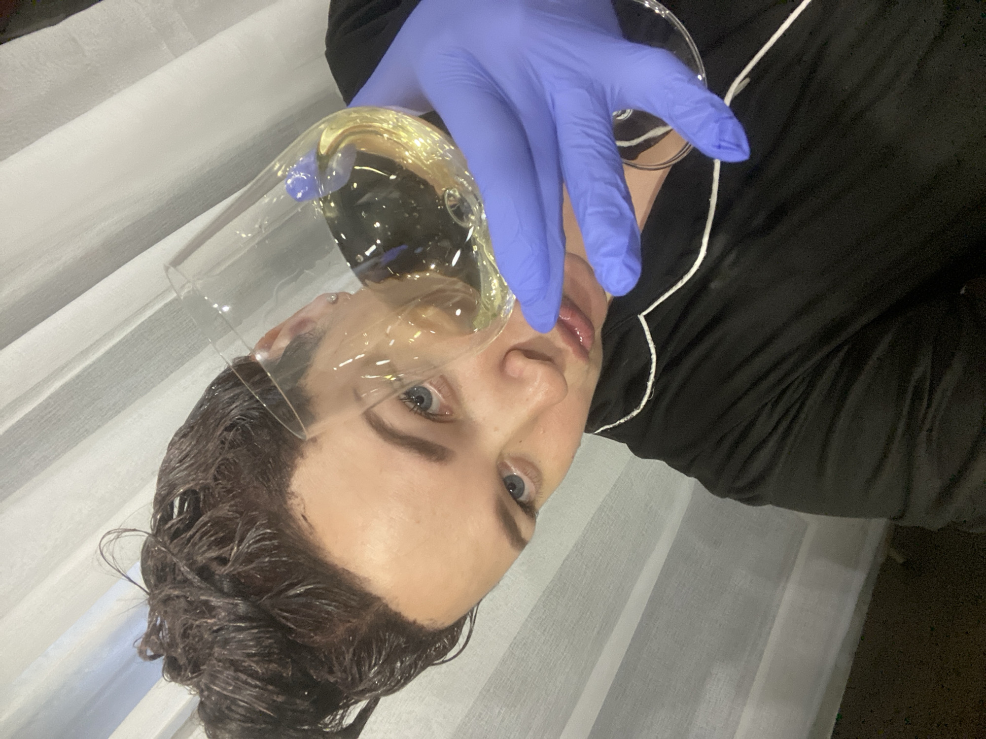 A person with blue eyes and dark, wet hair is positioned in front of a sheer, white curtain. The person is wearing a black top with white piping and holding a clear glass of yellow liquid with a gloved, blue hand near their face. The photograph appears to be taken indoors. There is no evidence to suggest the location of the city of Sochi, Russia.