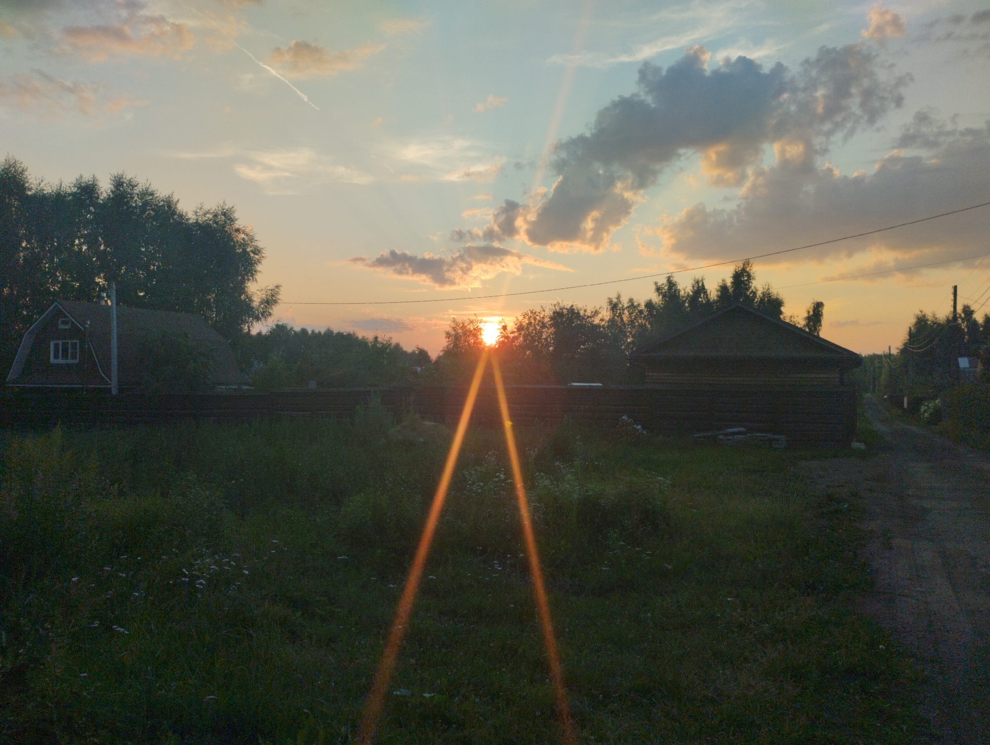 The image depicts a rural landscape at sunset, likely in or around Urkovo, Russia. The sky is a gradient of pale blue to orange, with scattered clouds tinged by the setting sun. Distinct sunbeams emanate from the horizon, creating a prominent lens flare that forms an inverted V-shape across the mid-ground. In the foreground, a grassy field with scattered white flowers stretches towards a dark wooden fence. To the left, a small, dark-colored building with a white-trimmed window is visible, partially obscured by trees. To the right of the fence, another dark, long, wooden structure is present, with a dirt path running alongside it. Dense trees line the background, creating a natural border between the cultivated land and the sky. No people or animals are present in the scene. The overall impression is one of quiet, natural beauty during the twilight hours.