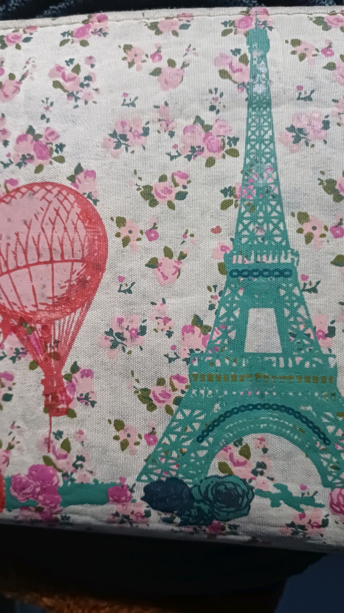 The image displays a close-up of a fabric with a floral pattern. Prominently featured are a stylized red hot air balloon on the left and a teal-colored Eiffel Tower on the right. The background is a light-colored fabric covered in small pink roses and green leaves. The fabric appears to be a decorative item, possibly a pillowcase or part of a bag. There are no people or animals visible. The image is a still photograph and does not provide any information about the city of Kowangan, Indonesia.